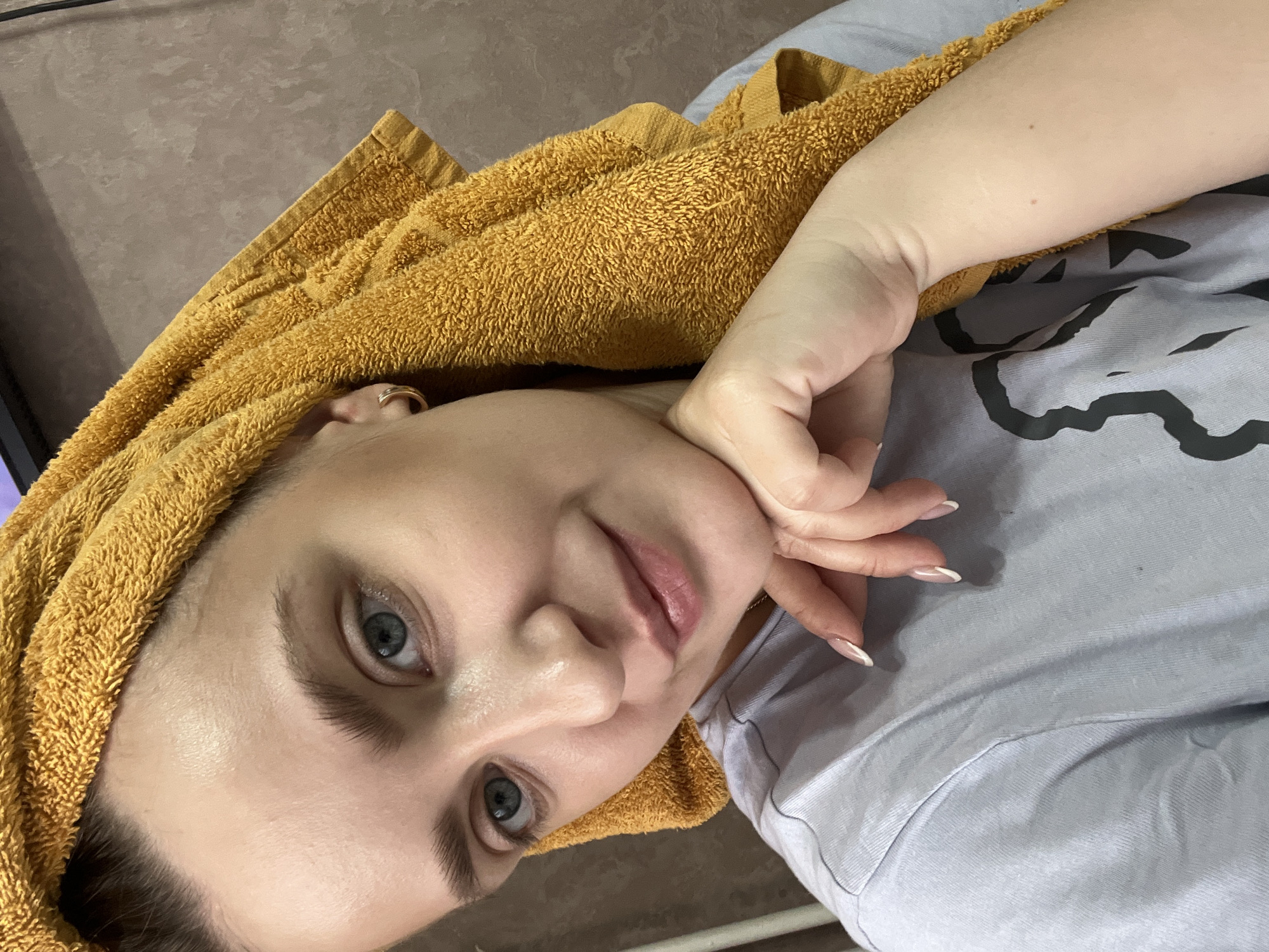 A woman with light skin and blue eyes is lying down, wearing a light grey t-shirt and an orange towel wrapped around her head. Her right hand is near her face. The location is Марковский, Russia. The background is a brown wall.