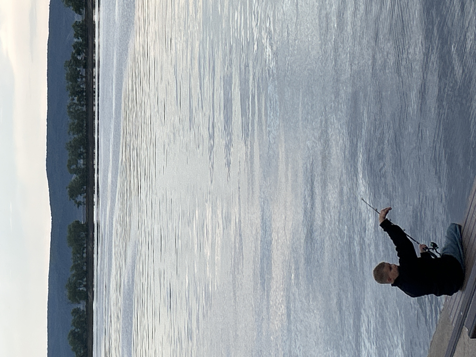 A person is fishing from a wooden pier. The person is wearing a black jacket and blue jeans and is holding a fishing rod. The person is sitting on the pier with their legs crossed. The fishing rod is extended over the water. The water is calm and reflects the sky. In the background, there are trees and a hill. The sky is overcast. The scene appears to be in Tolyatti, Russia. The time of day is likely late afternoon or early evening, given the soft light. There is no visible text in the image.