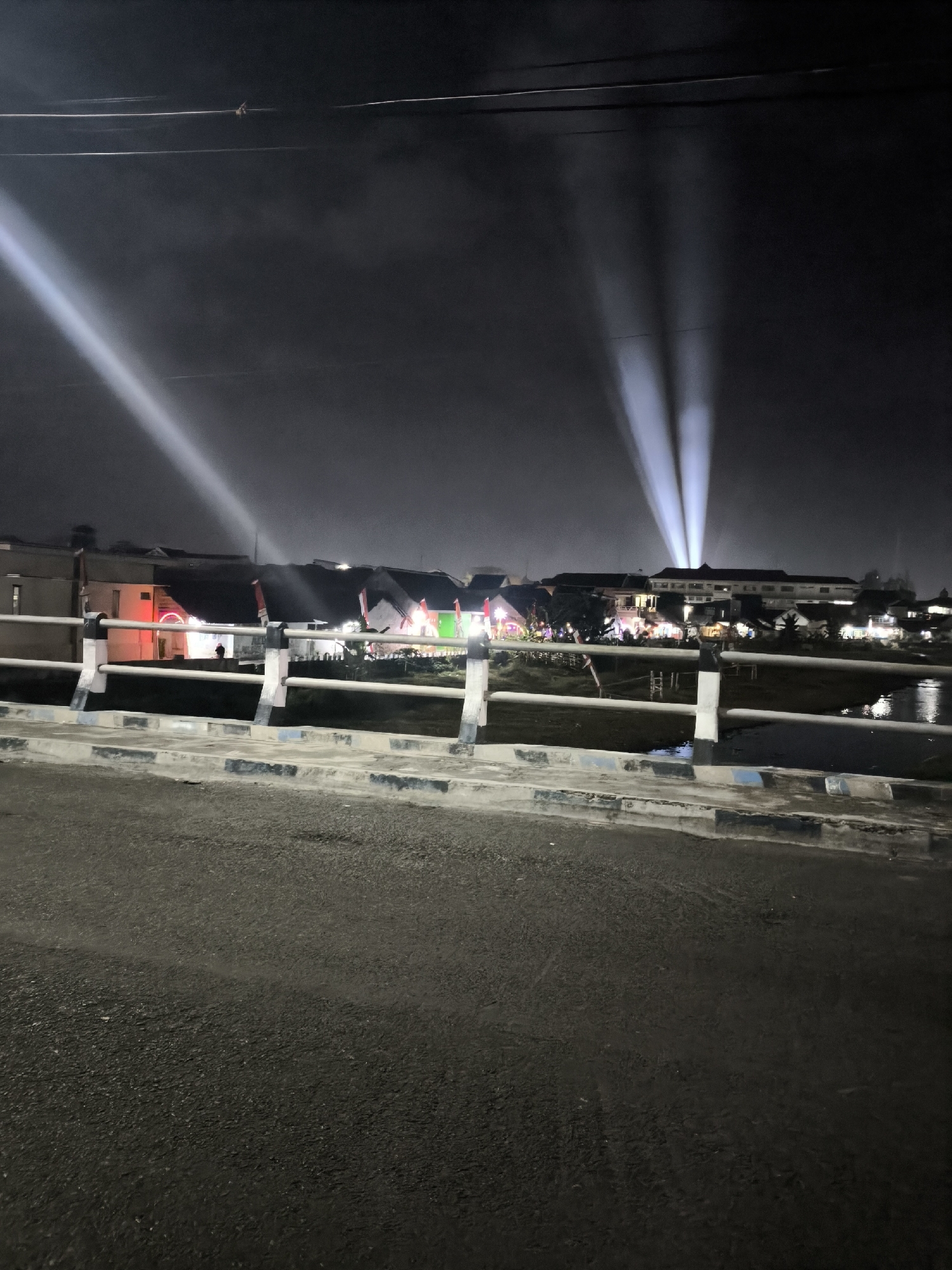 The image depicts a nighttime scene in Lumajang, Indonesia, likely taken from a bridge overlooking a body of water. The foreground is dominated by a dark asphalt road and a bridge railing. In the background, several buildings are visible, illuminated by lights and decorated with what appear to be Indonesian flags. Multiple spotlights are projecting beams of light upwards into the dark, hazy sky, creating a dramatic effect. The overall atmosphere suggests an event or celebration taking place in the city.