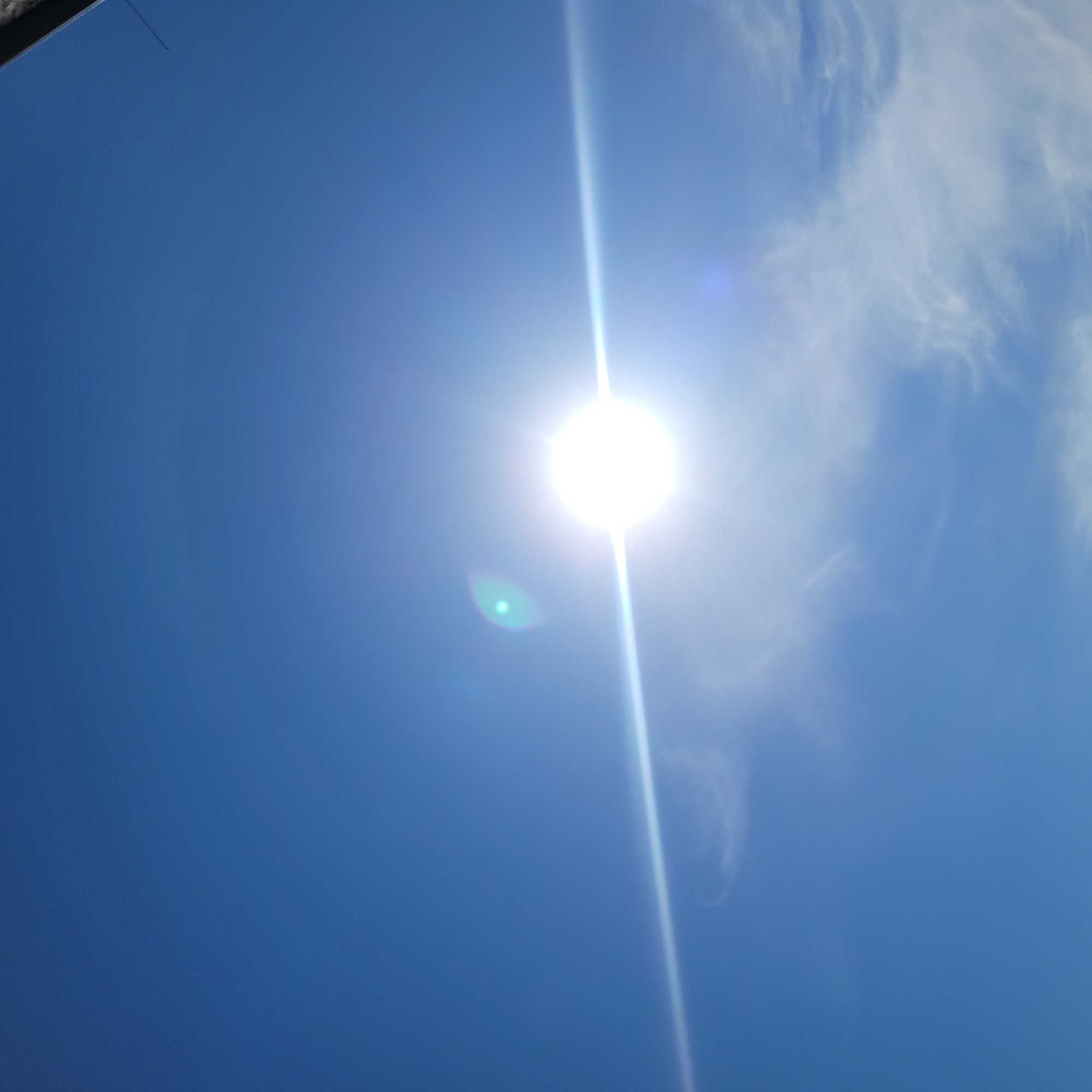 A bright sun with lens flare dominates the center of the image against a clear blue sky. A thin, wispy cloud formation is visible below and to the right of the sun. The upper right corner of the image shows a dark, possibly structural element, angled into the frame from the top. The environment appears to be outdoors, with the sky as the primary subject. No people or specific city landmarks in Jūrmala, Latvia are identifiable.