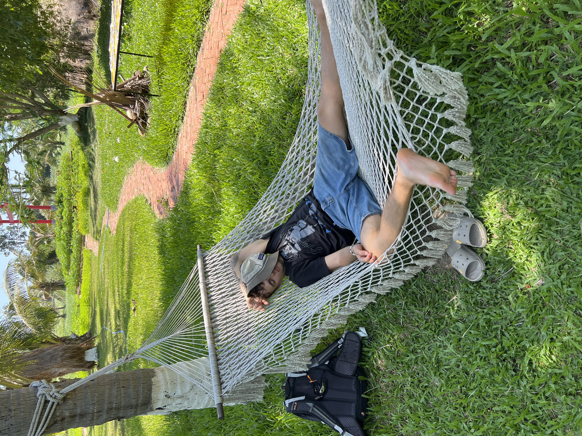 The image displays an outdoor scene in Nam Cam Ranh, Vietnam, under clear, sunny conditions. A single individual, appearing as a young adult male, is sleeping in a white net hammock. The hammock, featuring wooden spreader bars and decorative tassels, is secured to a thick-trunked palm tree, partially painted white, on the left side of the frame. The individual is dressed in a black t-shirt with a graphic print, blue denim shorts, and a light-colored bucket hat pulled over their face. Their right arm is bent with the hand under the hat, and a tattoo is visible on their left upper arm. A silver bracelet is on the left wrist. The person's bare feet are visible, and a pair of light grey slip-on clogs rests on the grass beneath the hammock. A black backpack with grey straps and orange accents is situated on the green grass adjacent to the hammock's left end.
The environment consists of a well-maintained green lawn. A winding red brick path cuts through the grass from the mid-left to the mid-right. To the right of the path, a small heap of dried vegetative matter is visible, alongside a dark-legged table supporting a flat, light-colored object. The background features various green trees and foliage, with a prominent red structure visible in the distance under a bright blue sky. The scene is brightly lit, casting distinct shadows.