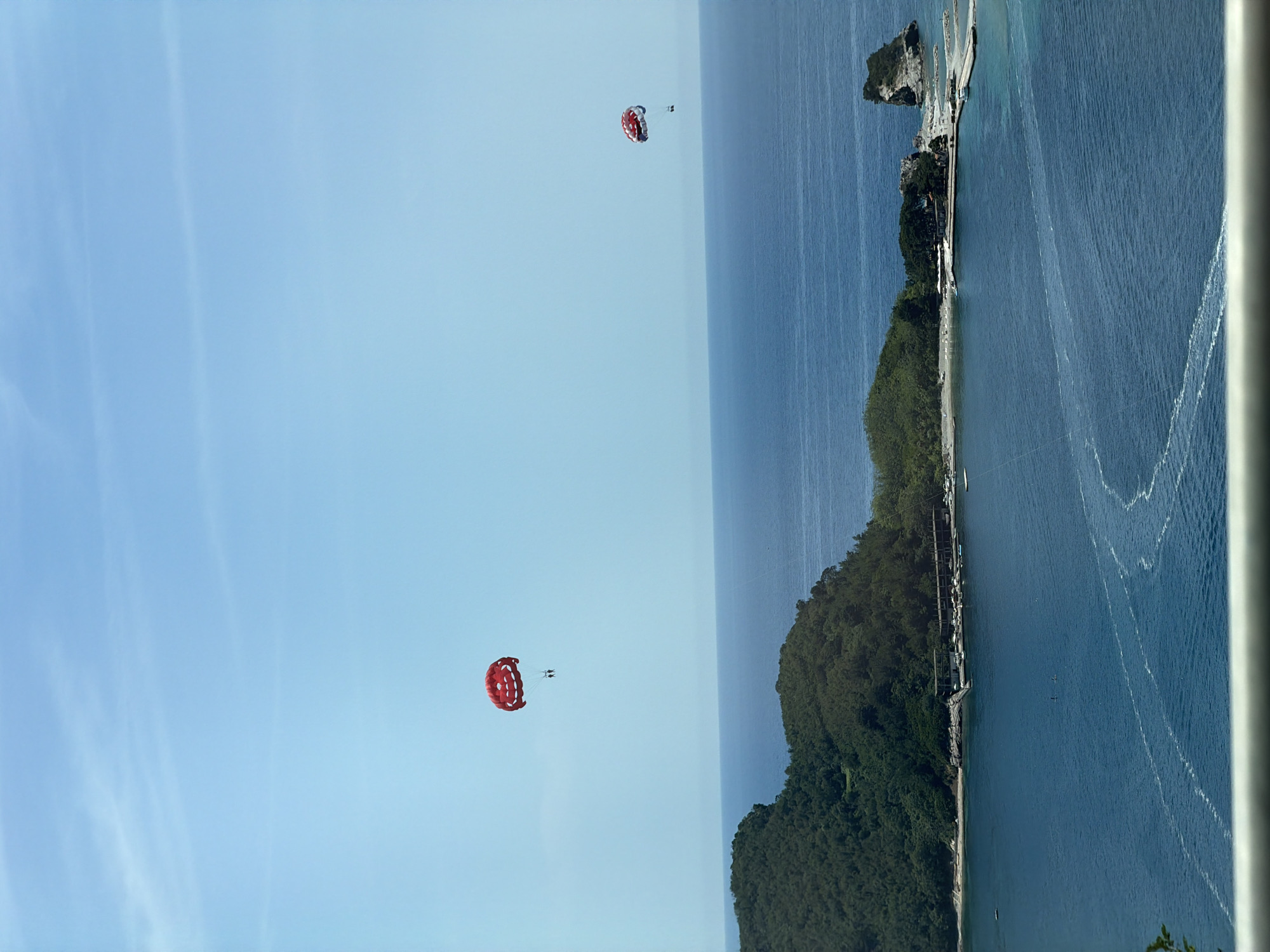 The image displays a panoramic coastal scene under a clear, light blue sky with faint cirrus clouds. The foreground features a light-colored railing. The middle ground is dominated by a vast expanse of calm, deep blue sea with visible white wake patterns. To the left, a large, verdant landmass, heavily forested with dark green trees, slopes down to the water. Along its base, a sandy beach is visible, transitioning into a developed area featuring buildings, a marina with numerous docked boats, and a pier extending into the water. Further to the right, a distinctive rocky island outcropping rises from the sea.
In the mid-left sky, a large, red and white parasail is prominently featured, with two individuals suspended beneath it. To the right, further in the distance, a smaller, similarly colored red and white parasail is airborne, with a single individual beneath it. Subtle ripples and reflections are visible on the water's surface, particularly near the land. The scene depicts a bright, sunny day over the Adriatic Sea, characteristic of the coastal area in Boreti, Montenegro, with recreational watersports in progress.