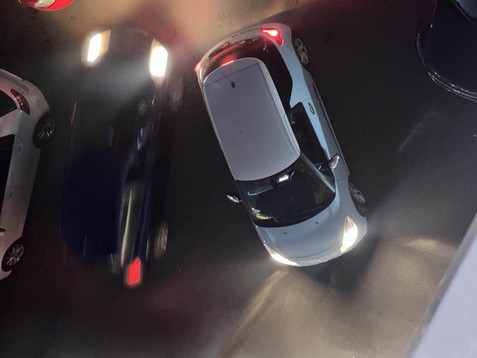 The image shows an aerial view of a street scene at night, likely in Belgrade, Serbia, based on the car models. The scene includes several cars parked closely together. A white or light gray car is prominently positioned in the right center of the frame, with its headlights illuminated. Another dark-colored vehicle is visible to the left, with headlights reflecting brightly. Additional cars are partially visible at the edges of the frame. The environment appears to be a dark, paved road surface. The lighting in the scene is a mix of artificial light from the car headlights and taillights, creating strong highlights and shadows. The overall impression is of a congested urban area at night.