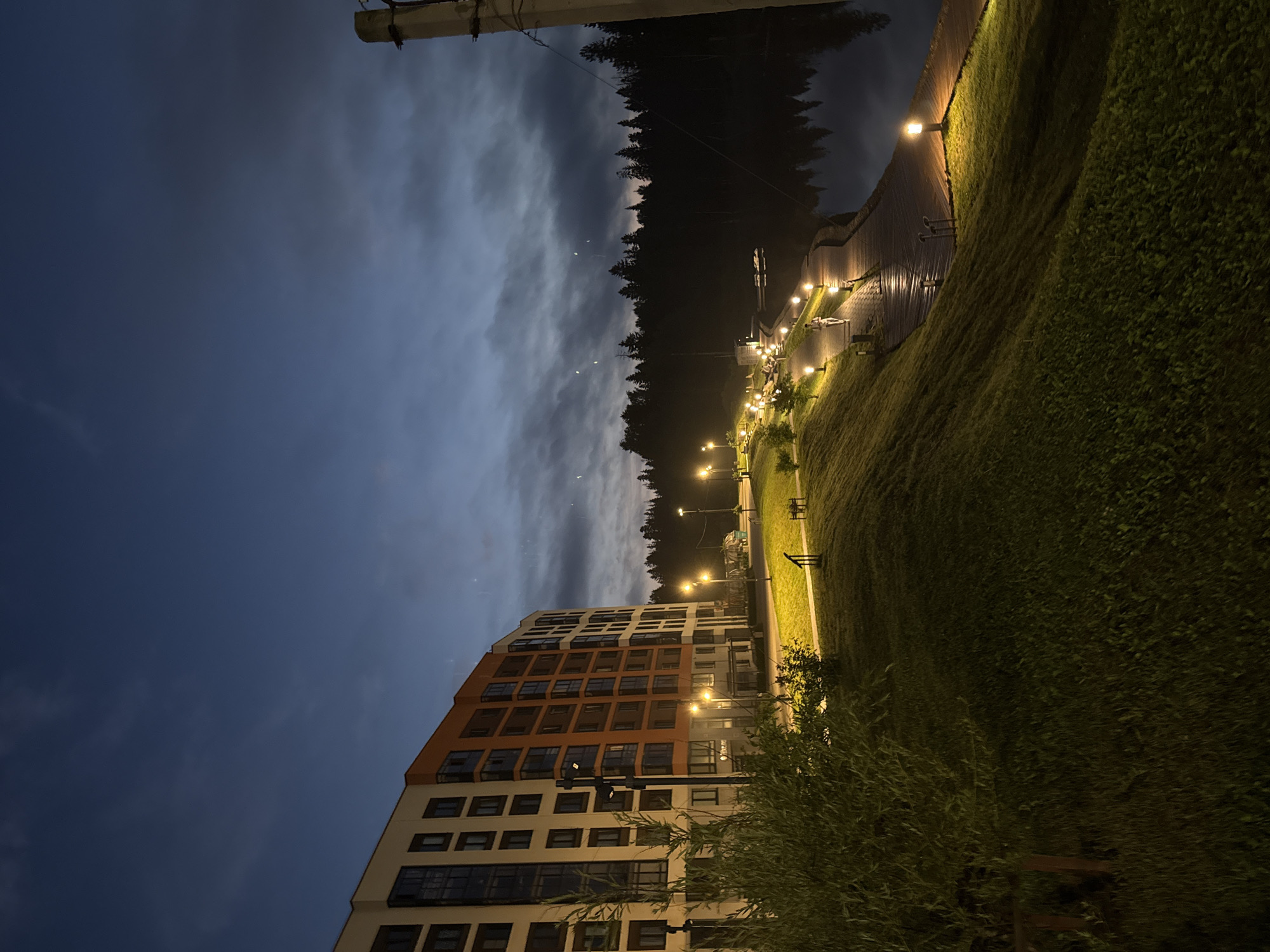 The image depicts an evening scene in Izhevsk, Russia, likely near a body of water. A modern, multi-story apartment building with white and orange facades dominates the left side of the frame. In front of the building, a grassy slope leads down to a paved walkway lined with lampposts casting a warm glow. The walkway follows the edge of a dark body of water, reflecting the lights and the silhouetted treeline on the opposite shore. The sky is a deep twilight blue with scattered clouds, and a few stars are visible. The overall atmosphere is tranquil, suggesting a residential area with waterfront amenities.