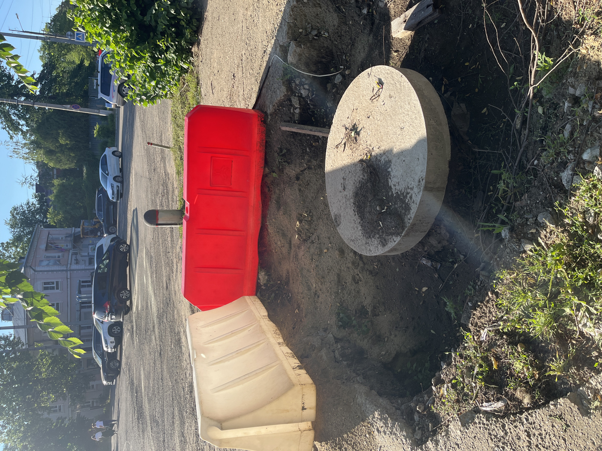 A construction site is visible in an urban environment, possibly in Krasnodar, Russia, with a large excavated pit in the foreground. The pit is partially covered by a white, plastic, water-filled barrier on the left and a red, plastic, water-filled barrier on the right. A large, circular concrete slab rests on the edge of the pit, partially obscuring it. The ground around the pit is a mixture of dirt, gravel, and some greenery. In the background, several cars are parked on a paved surface, and buildings and trees are visible. A few people are walking in the background near the parked cars. The scene appears to be during daylight hours, with sunlight casting shadows.