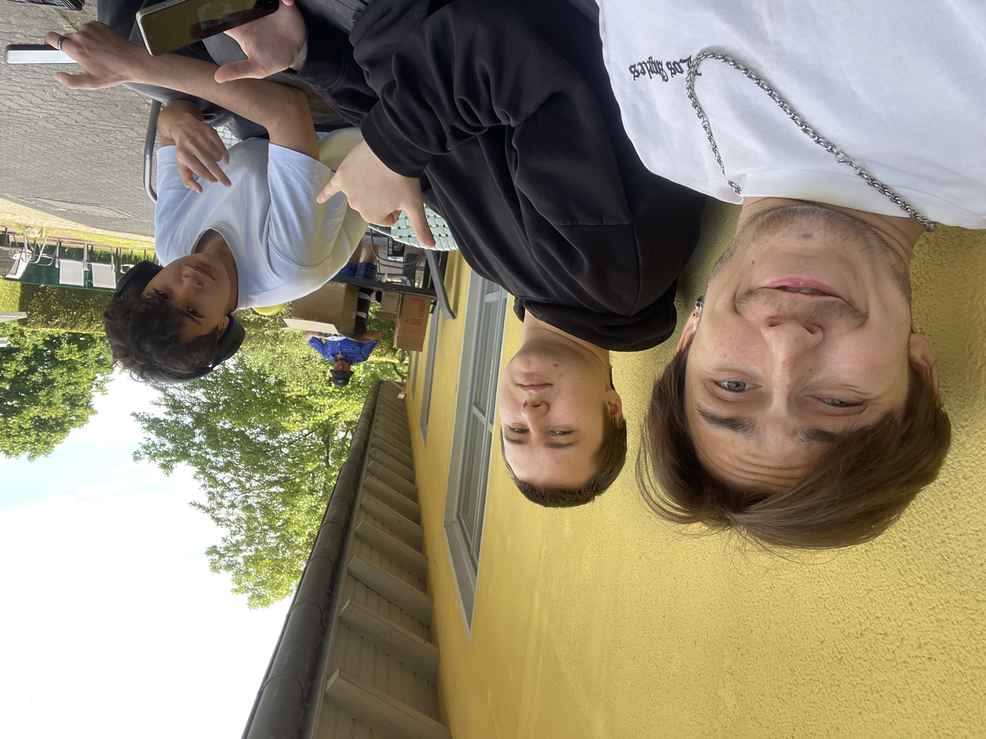 Three young men are posing for a photo outdoors. The man on the left is wearing a white t-shirt and headphones. The man in the middle is wearing a black hoodie. The man on the right is wearing a white t-shirt with a silver chain. They are sitting at a table on a paved area. Behind them is a yellow building with a window. In the background, there are trees and a person wearing a blue shirt and a hat. The location is Wienhausen, Germany.
