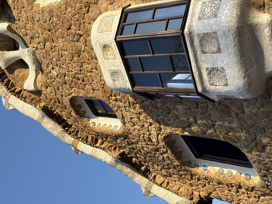 An upward-angled view captures a section of a highly textured and artistically detailed building facade against a clear blue sky. The structure is identified as one of the gatehouses at Park Güell in Barcelona, Spain. The primary wall surface consists of irregularly shaped, rough-hewn, medium-brown stone blocks, showing deep shadows that emphasize their uneven texture. Architectural elements such as the undulating roofline, window frames, and a prominent bay window are adorned with "trencadís" (broken ceramic tile mosaic).
The mosaic work is predominantly white, with decorative patterns in blue, yellow, red, and green accentuating the curves and framing the windows. One irregularly shaped window opening is visible on the upper left, framed by mosaic patterns featuring concentric circles. Another similar window is present on the mid-left. A multi-paned bay window, situated in the lower right and projecting from the facade, features dark brown frames and clear glass reflecting the blue sky and sparse tree branches. The base and top of this bay window are covered in white trencadís, incorporating four distinct square decorative tiles with intricate geometric and floral patterns in blues, yellows, and oranges. Sunlight from the upper left casts sharp shadows across the facade, highlighting the three-dimensional quality of the stone and mosaic work. No individuals or additional objects are present in the scene.