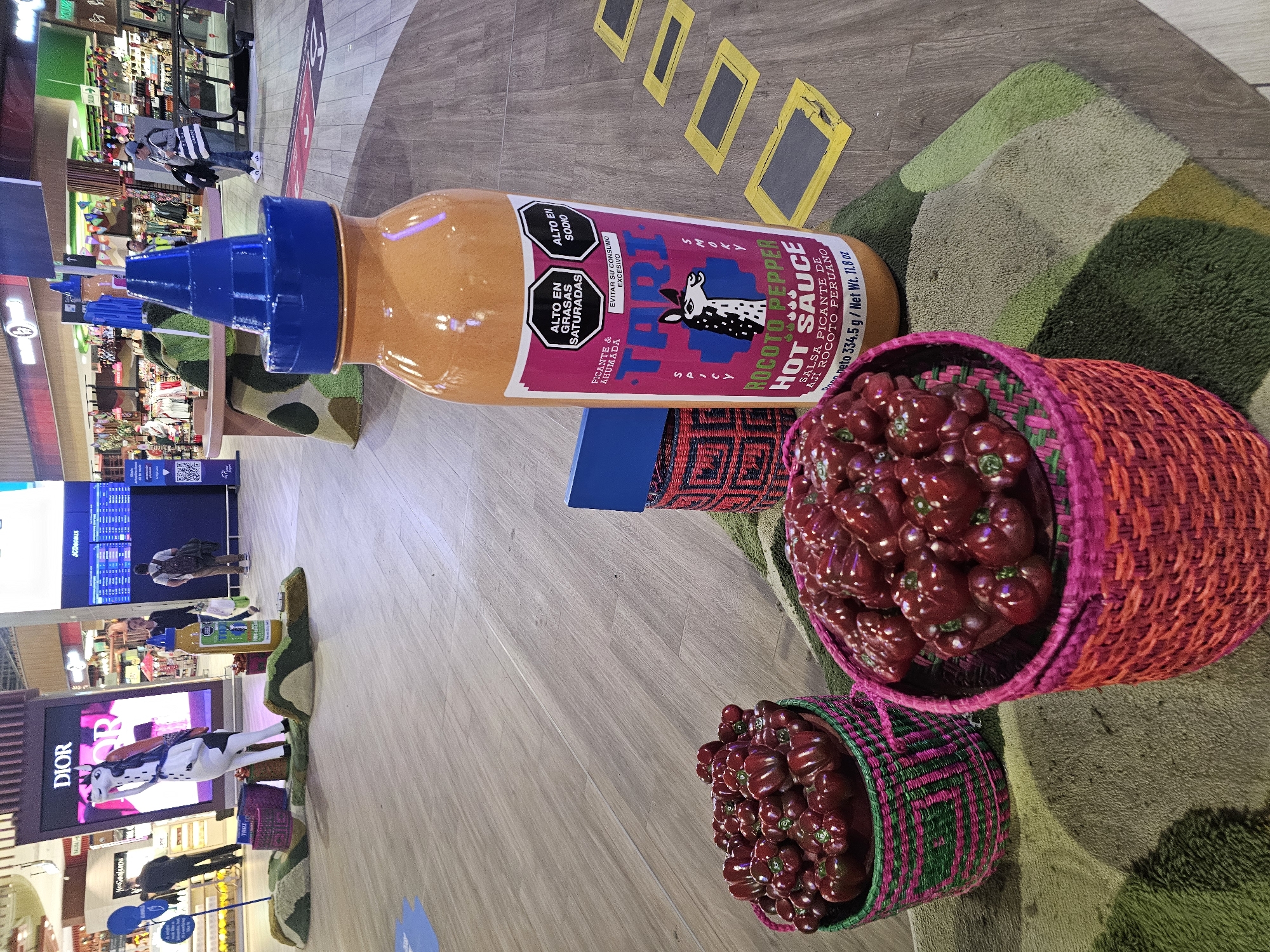 Here is a detailed description of the image:
**Scene Overview:**
The image appears to be taken inside a commercial space, possibly an airport or shopping mall in Callao, Peru. It shows a large-scale art installation or display featuring Peruvian themes.
**Objects:**
* A massive replica of a bottle of "TRI" Rocoto Pepper Hot Sauce is the most prominent object.
* Several oversized baskets are filled with what appears to be artificial Rocoto peppers.
* The floor has a light wood-grain pattern.
* There is a series of yellow rectangular shapes on the floor, perhaps decorative or directional.
* Several commercial storefronts, including a "DIOR" boutique, are visible in the background.
* Electronic flight information boards are mounted on a wall in the background.
**People:**
* Several people are visible in the background, including shoppers or travelers.
**Environment:**
* The space is indoors and well-lit.
* The background suggests a bustling commercial environment.
**Overall Impression:**
The image captures a visually striking display promoting Peruvian culture, potentially related to food products or tourism. The use of oversized items creates a sense of novelty.