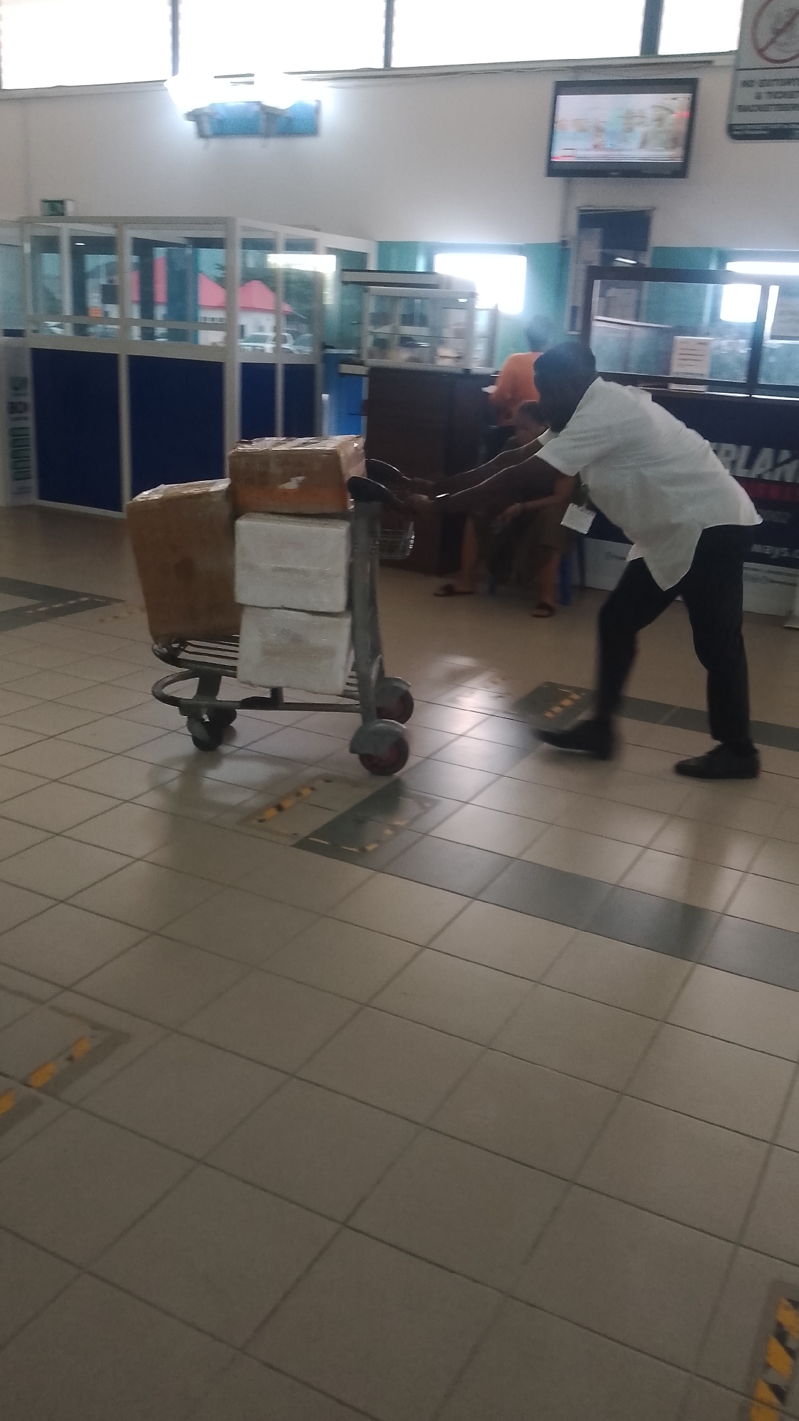 A person is pushing a luggage cart with boxes in what appears to be an airport terminal. The person is wearing a white shirt and dark pants. In the background are other people sitting and standing. A television is mounted on the wall. The floor is tiled. The city of Orerokpe, Nigeria, is mentioned in the description request, but this cannot be verified from the image.