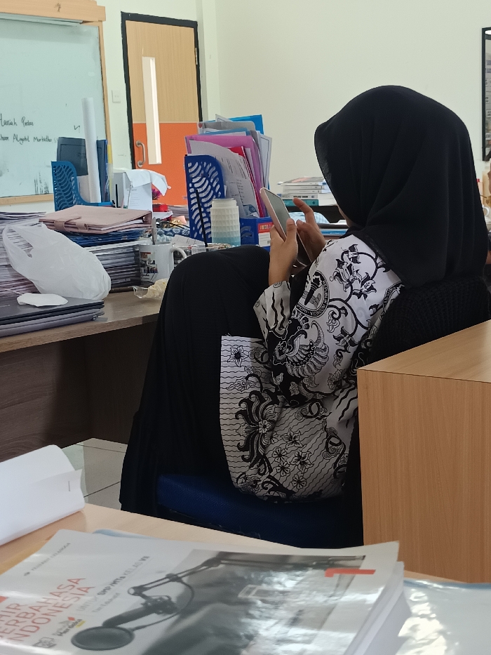 A woman in a black hijab and patterned top sits at a desk, looking at her phone. The desk is cluttered with papers, books, and various office supplies, including a white board. A doorway is visible in the background. More books are in the foreground on another desk.
