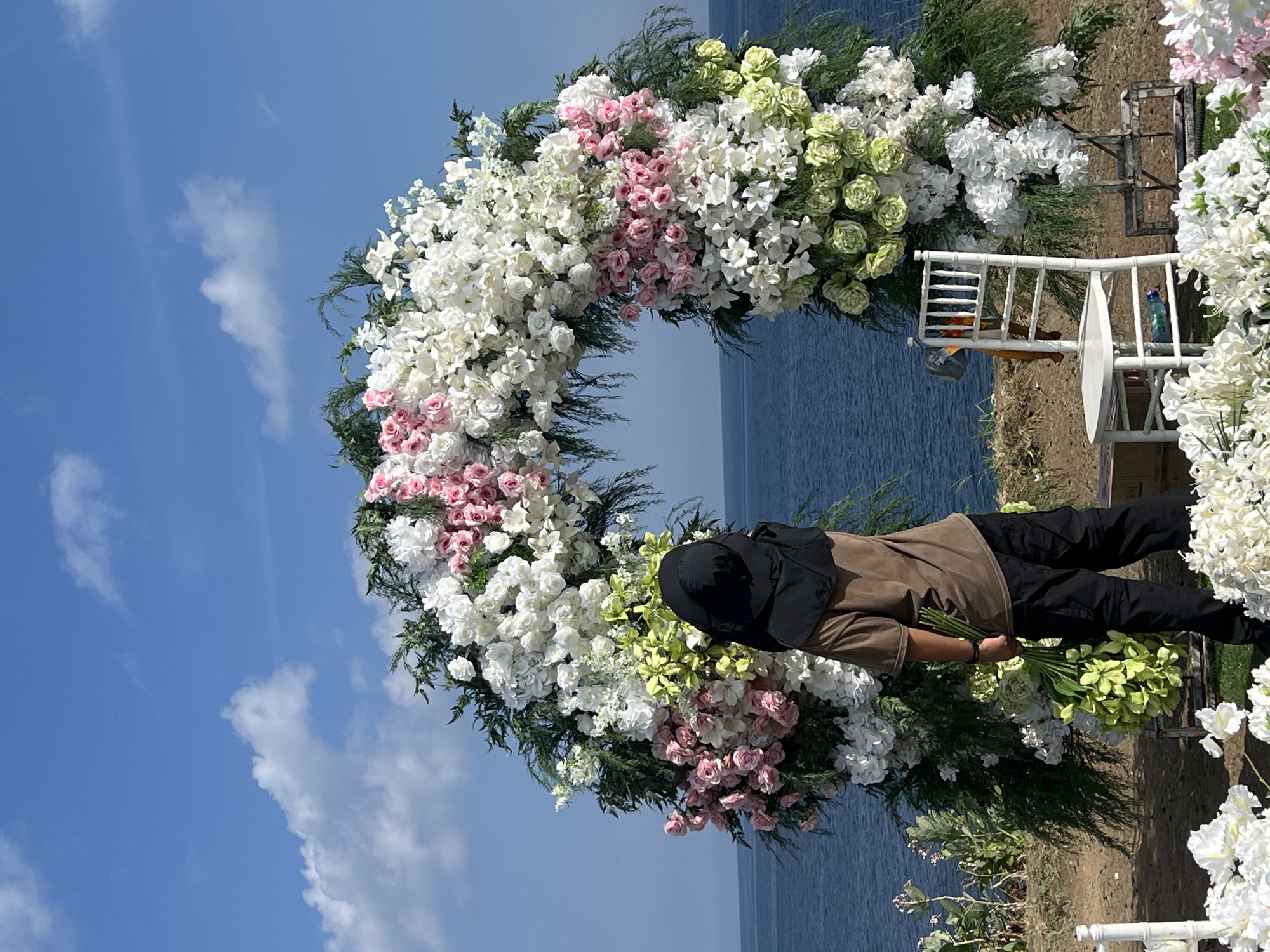 A vibrant outdoor scene in Kuta Selatan, Indonesia, captures the preparation for a special event by the ocean. Under a bright blue sky with scattered white clouds, a large, circular floral archway stands prominently, densely decorated with a mix of white, light pink, and light green flowers, complemented by dark green foliage. The white and light green blooms appear to be hydrangeas, stock, and similar dense varieties, while the pink flowers are smaller roses.
On the right side of the archway, a person wearing a black wide-brimmed hat, a tan short-sleeved top, and black trousers is focused on arranging flowers, holding a fresh bouquet of light green blooms. Two white ornate chairs are positioned near the arch's base; one holds a blue water bottle. The deep blue ocean forms a scenic backdrop, meeting the horizon. The bright daylight indicates a mid-morning or afternoon setting, conveying a sense of anticipation for an elegant coastal ceremony. No visible text is present.