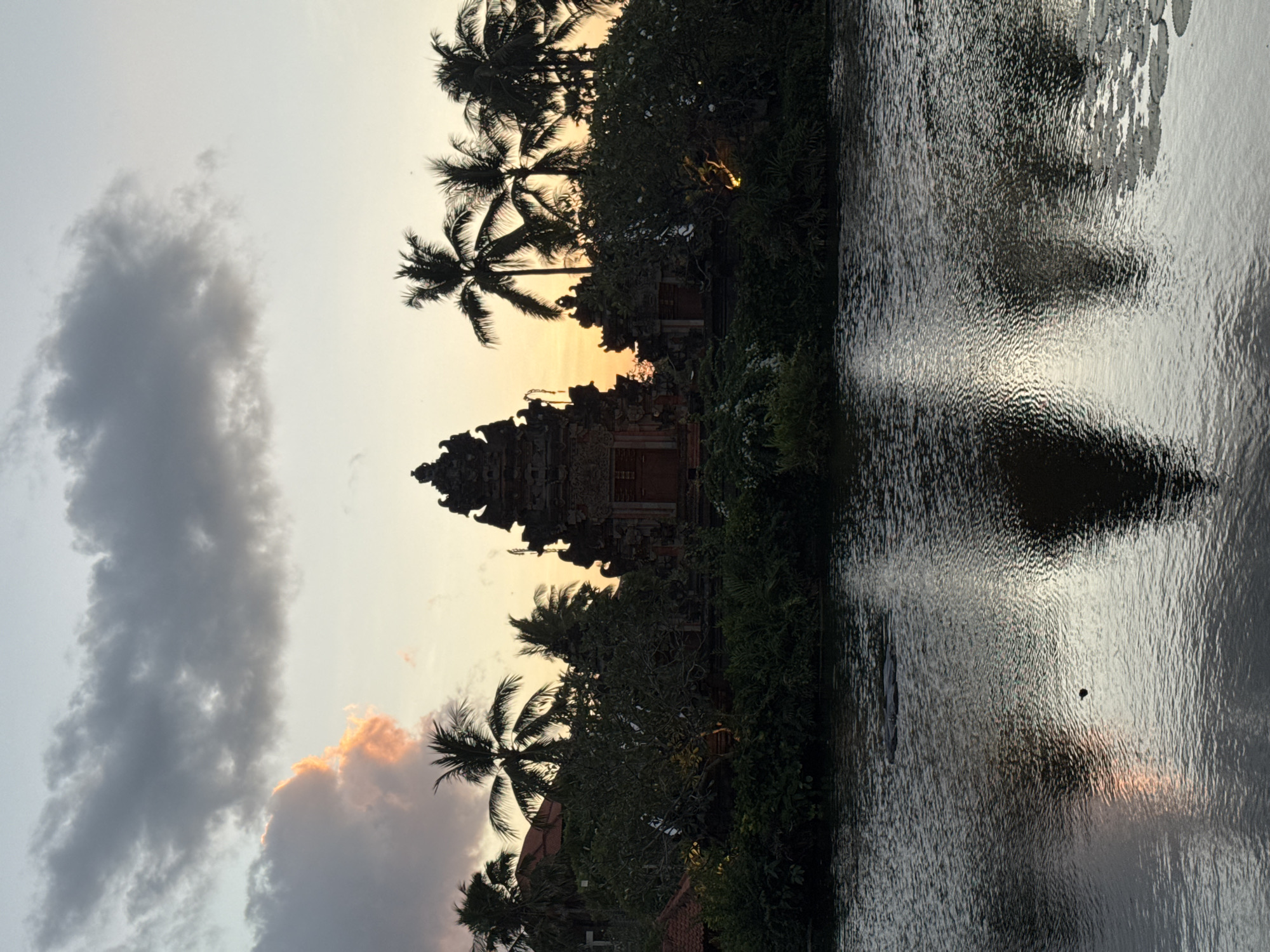 The image depicts a tranquil scene at dusk in Kuta Selatan, Indonesia, with a prominent ancient temple structure silhouetted against the colorful sky. The temple, characterized by its tiered roof and intricate carvings, is surrounded by lush greenery and towering palm trees. The foreground features a calm body of water, its surface reflecting the twilight hues of the sky and the temple's silhouette. The water is disturbed by gentle ripples, suggesting a serene atmosphere. Scattered across the water's surface are a few lily pads. The sky displays a gradient of soft oranges, yellows, and blues, with dramatic clouds adding depth and texture to the composition. The overall impression is one of peacefulness and cultural immersion, typical of the tropical landscapes found in Kuta Selatan.