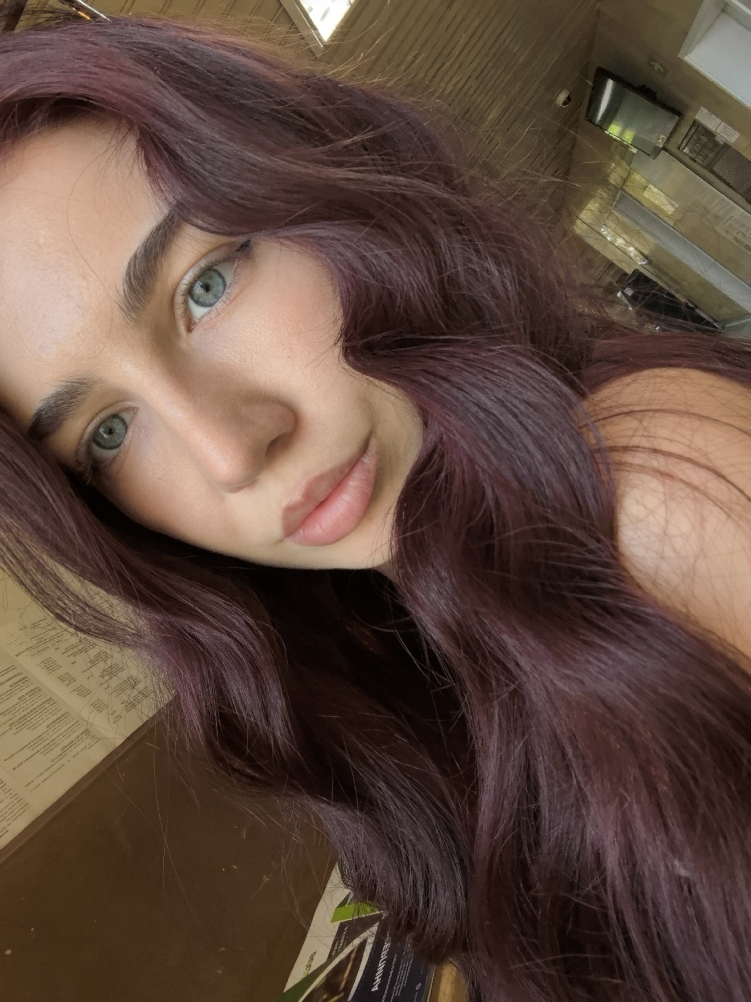 A close-up, slightly angled selfie of a young woman with long, wavy auburn-colored hair. Her eyes are light green. Her skin is light-toned. She has a light application of makeup. Behind her, out of focus, is a room with a television mounted on the wall. There is also a counter. The image does not contain enough detail to determine if the location is in Bogomilovo, Bulgaria.