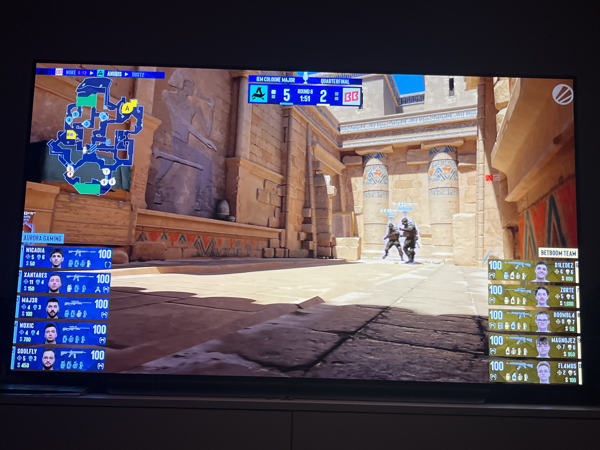 This is a television screen displaying a professional esports match, likely Counter-Strike: Global Offensive, given the user interface elements. The game is taking place on a map that appears to be "Anubis," as indicated by the map selection bar at the top. The score is 5-2 in favor of the team labeled "BB." The screen shows in-game player information, including player names, kill/death/assist counts, and money. Two players are visible in the game environment, engaged in combat. The scene is set in a visually elaborate virtual environment with Egyptian architectural influences. The screen itself is mounted on furniture, suggesting a home viewing setting. There is no indication of the city of Varna, Bulgaria in this image.