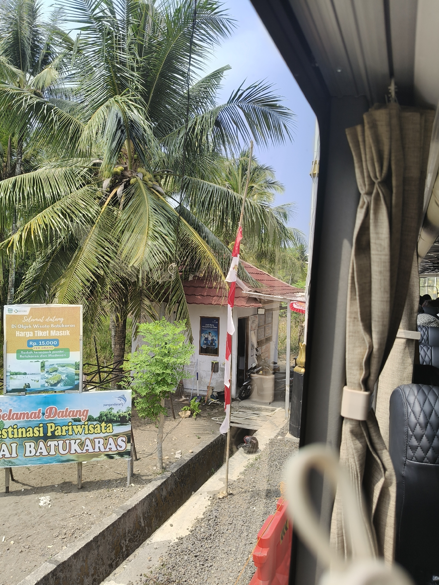 The image captures a view from inside a vehicle, looking out at a roadside scene in Desa Batukaras, Indonesia. Dominating the scene is a cluster of lush green coconut trees, their fronds reaching towards a bright, clear sky. To the left, a large sign displays promotional information for a tourist destination, with text in Indonesian that translates to "Welcome to the Batukaras Tourist Attraction." Below this, it lists the entrance ticket price as Rp. 15,000 per person, and states that it includes access to Batukaras and Madadari beaches. Images on the sign depict scenic coastal areas.

In the mid-ground, there is a small, simple building with a red-tiled roof, possibly a ticket booth or information center for the attraction. A red and white Indonesian flag flies from a pole in front of the building. A small, leafy tree stands to the right of the sign, and a motorcycle is parked near the building. The ground is covered with gravel and dirt, and a concrete drainage ditch runs along the foreground. On the right side of the image, a beige curtain from the vehicle is partially visible, along with the edge of a dark-colored, upholstered seat. The overall impression is one of a tropical tourist location on a sunny day.