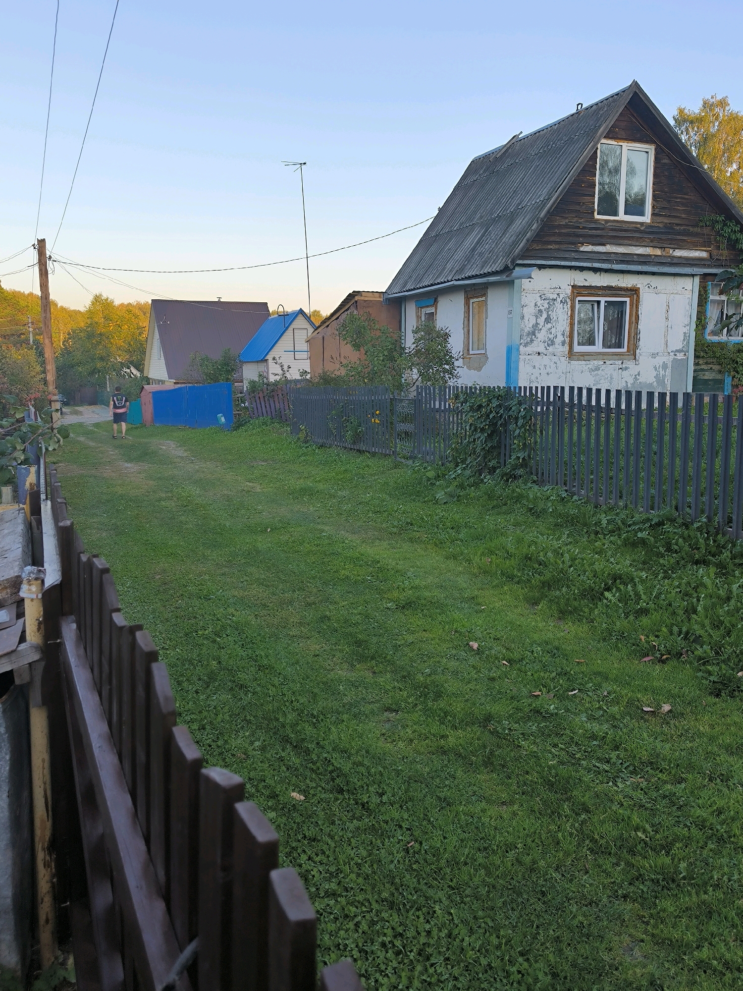 The image depicts a rural residential scene in Maiskii, Russia. In the foreground, a dark brown wooden fence runs along the left side of a grassy path. The path leads into the distance towards several houses. A young person wearing a dark t-shirt and shorts is walking away from the camera down the path. To the right of the path, a dark grey vertical slatted fence encloses a property with a weathered, two-story house. The house has a dark grey roof, wooden upper walls, and a white, peeling lower facade with blue trim around some windows. Further down the path, other houses are visible, including one with a blue roof and another with a brown roof. The environment is characterized by lush green grass, trees with autumn foliage, and a clear, pale blue sky, suggesting it is late afternoon or early evening. Power lines and a utility pole are also present.