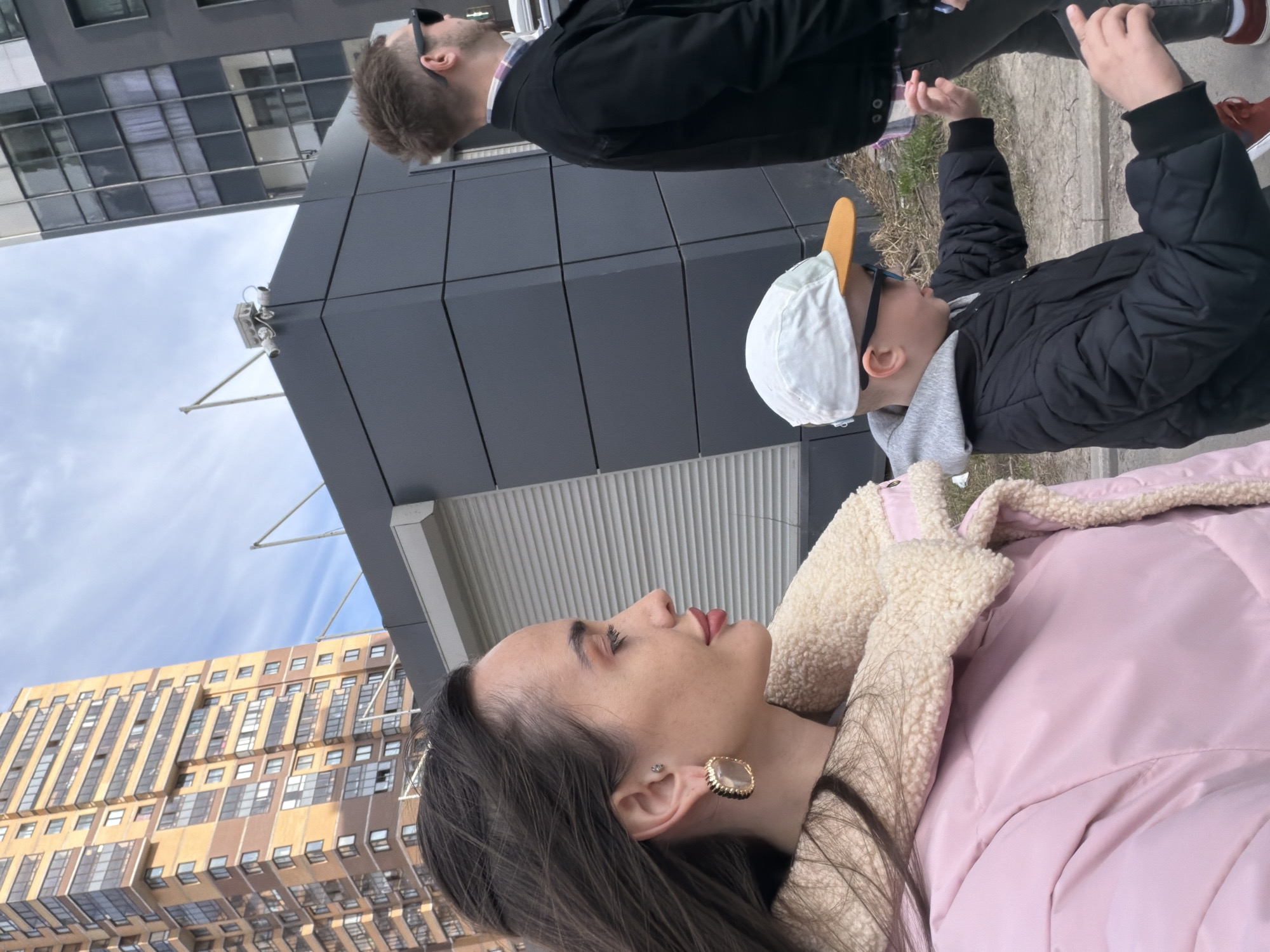 The image shows a medium shot of three people in an urban environment, likely Saint Petersburg, Russia. A woman with dark brown hair, wearing a pink winter jacket with a cream-colored fleece collar, is in the foreground. Behind her, a boy wearing a white baseball cap with an orange bill, sunglasses, and a black puffer jacket is looking up and slightly to the right. Further back, a man with sunglasses, wearing a black jacket and jeans, is also looking in the same direction as the boy. The background includes a modern building with gray paneling, another taller building with numerous windows, and a blue sky. The people appear to be walking or standing on a sidewalk or paved area.