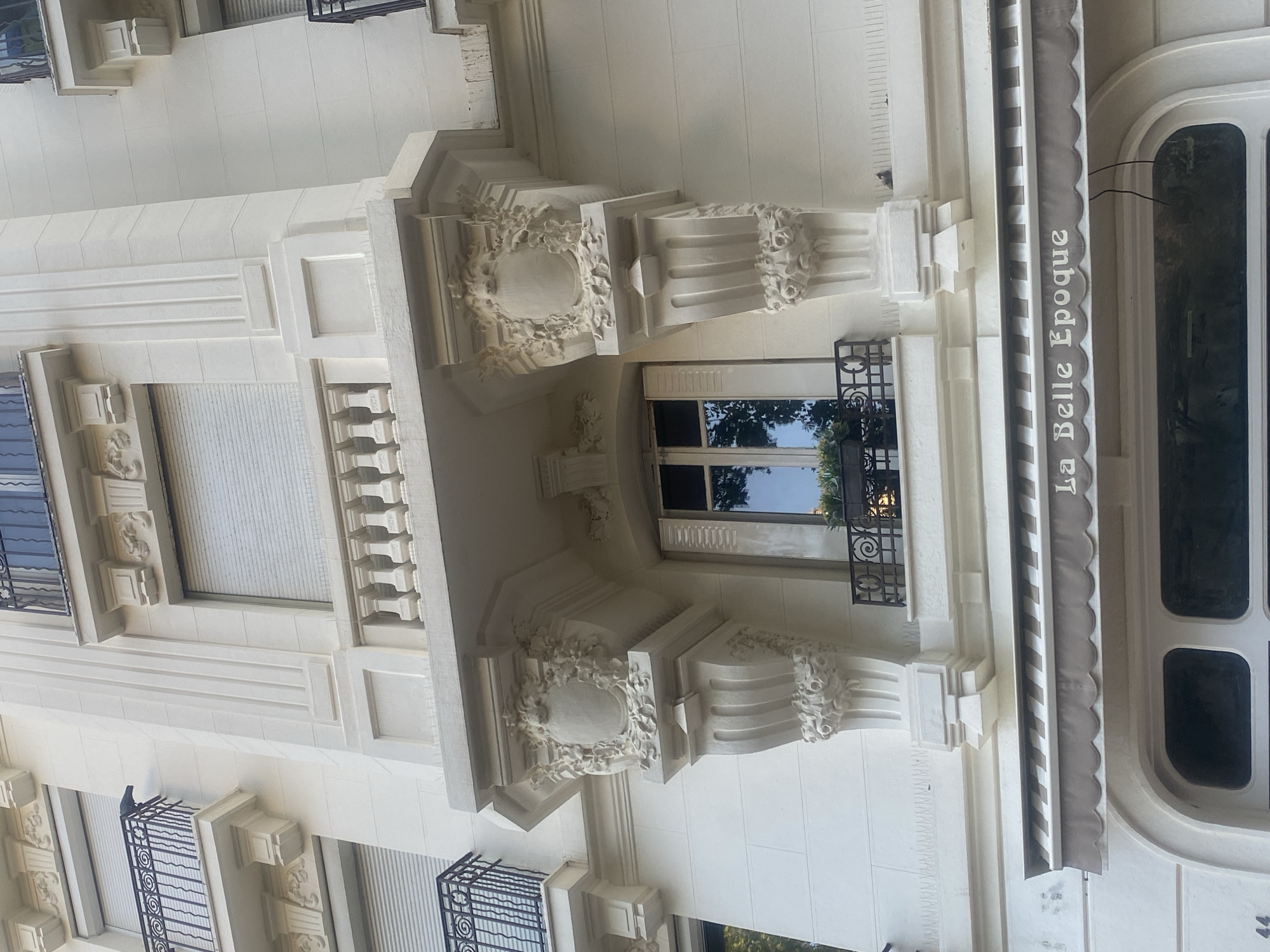 The photograph captures the facade of a building in Antibes, France, likely a shop or restaurant, given the awning that reads "La Belle Epoque". The building's exterior is an ornate, creamy white, adorned with classical architectural elements. A central balcony, supported by intricately carved corbels featuring floral motifs and what appears to be a crest, juts out from the building's second story. Below this balcony, a window is visible with a small railing in front of it, containing a planter with greenery. The building also features multiple windows with closed blinds and decorative railings on other balconies. The overall impression is one of historic elegance.