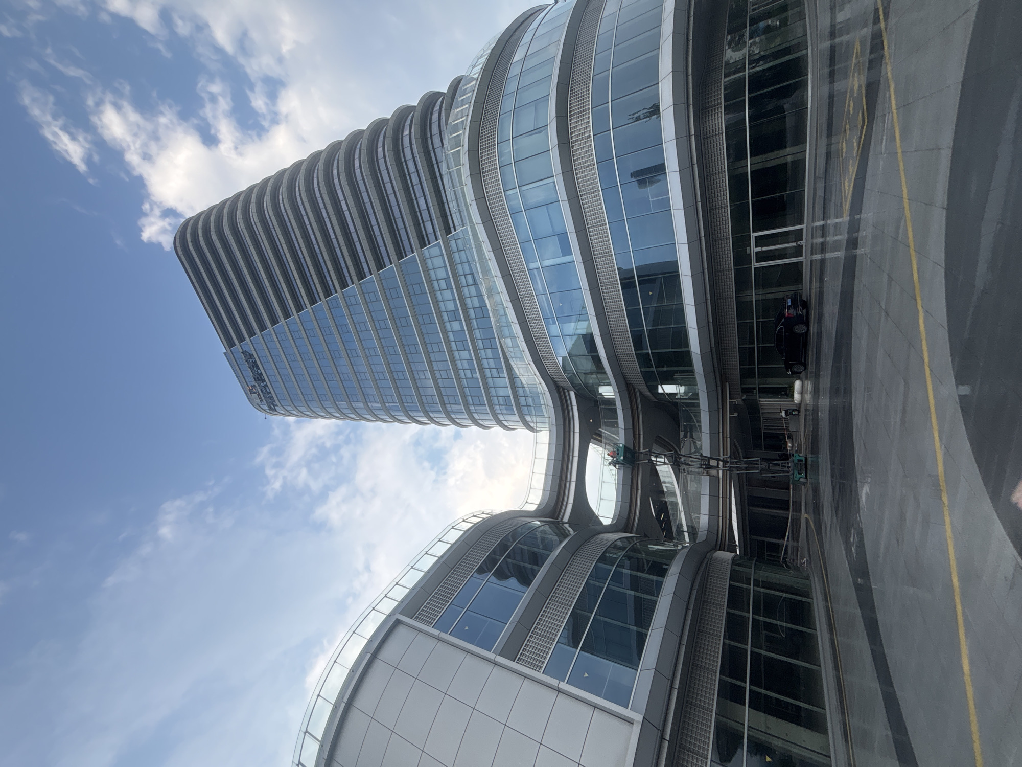 The image captures a modern architectural complex in Suzhou Industrial Park, China, characterized by its sleek, curved buildings and extensive glass facades. A tall, multi-story building with horizontal banding and a prominent slanted roof dominates the right side of the frame, featuring a glass exterior that reflects the sky. To the left, a lower, curved structure with similar glass windows and metal paneling connects to the taller building via elevated walkways.
In the foreground, a wet, paved area with yellow markings suggests a plaza or parking space, reflecting the surrounding architecture. A dark-colored sedan is parked near the entrance of the curved building on the right. A mobile elevating work platform (MEWP) is positioned near the center of the image, with two individuals visible in the basket at a mid-level height, seemingly engaged in maintenance or cleaning activities on the exterior of the connecting walkways. The sky above is partially cloudy, with a mix of blue sky and white clouds, indicating daylight conditions. The overall scene portrays a contemporary urban environment with advanced architectural design.