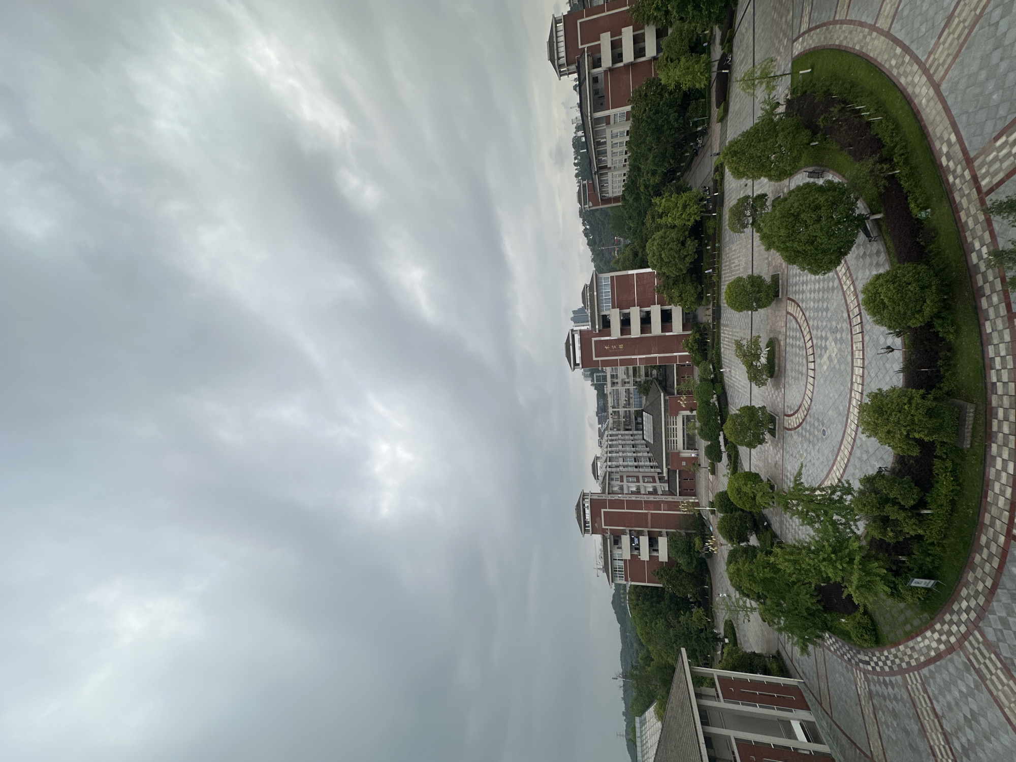 The image shows an aerial view of an outdoor scene in Chongqing High-tech Zone, China, under an overcast sky. Several multi-story red-brick buildings are interspersed with lush green trees. A circular paved area with concentric patterned paths is visible, featuring manicured bushes and trees. The overall atmosphere is urban, with a focus on landscaped public space. No people are discernible in the image.