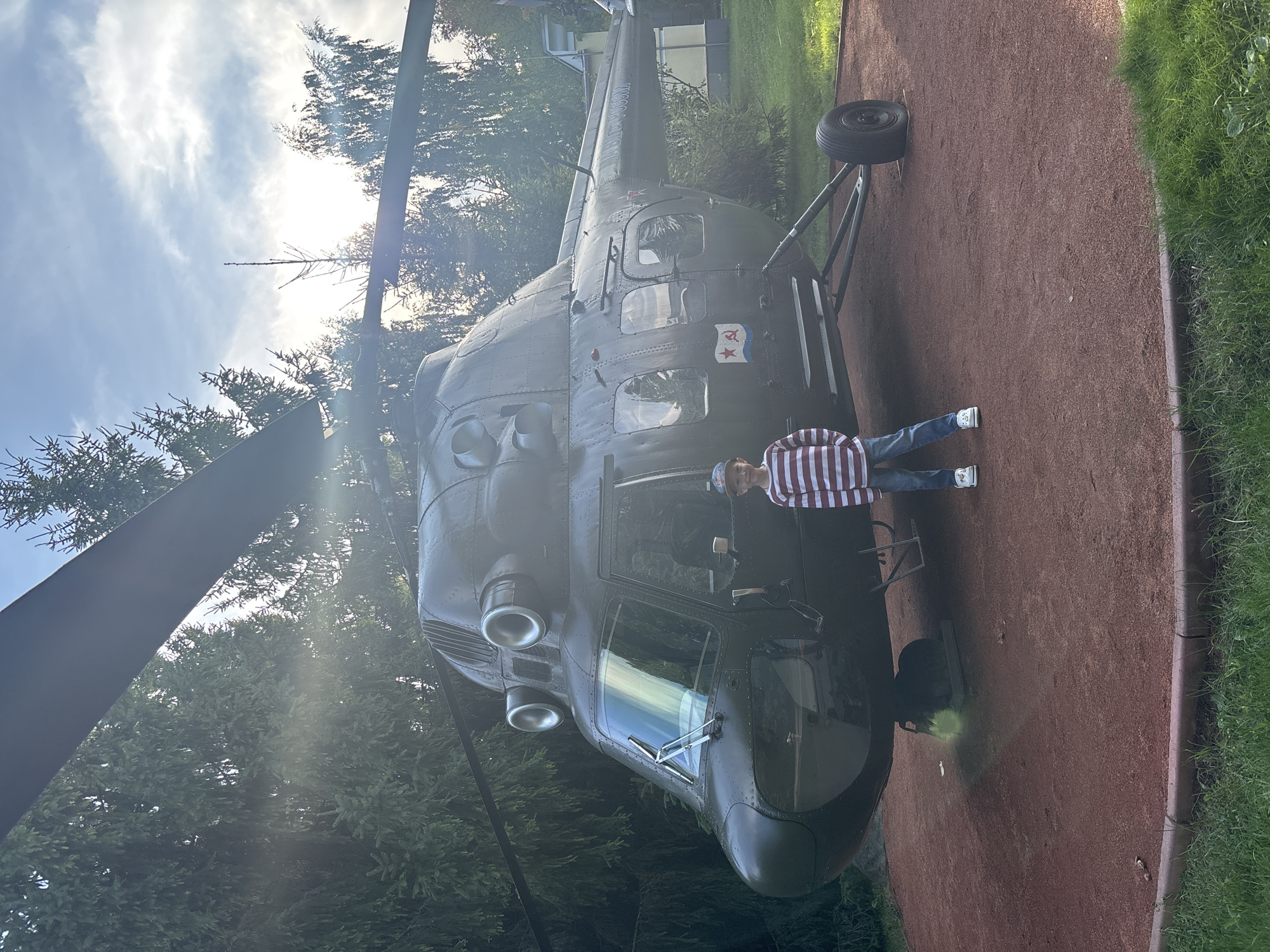 A young boy stands in front of a parked dark green helicopter on a reddish-brown gravel surface surrounded by green grass. The helicopter is large with visible rotor blades overhead and a prominent cockpit with windshield wipers. The boy, wearing a striped long-sleeved shirt, blue jeans, a baseball cap, and white sneakers, is positioned to the right of the helicopter's nose. The background shows lush green trees and a bright sky with sunlight filtering through the foliage. A small red insignia with a hammer and sickle is visible on the side of the helicopter. The scene is likely outdoors, possibly in a park or museum exhibiting military equipment. The image does not provide explicit details about the city of Рождествено, Russia, beyond its potential inclusion as a location.