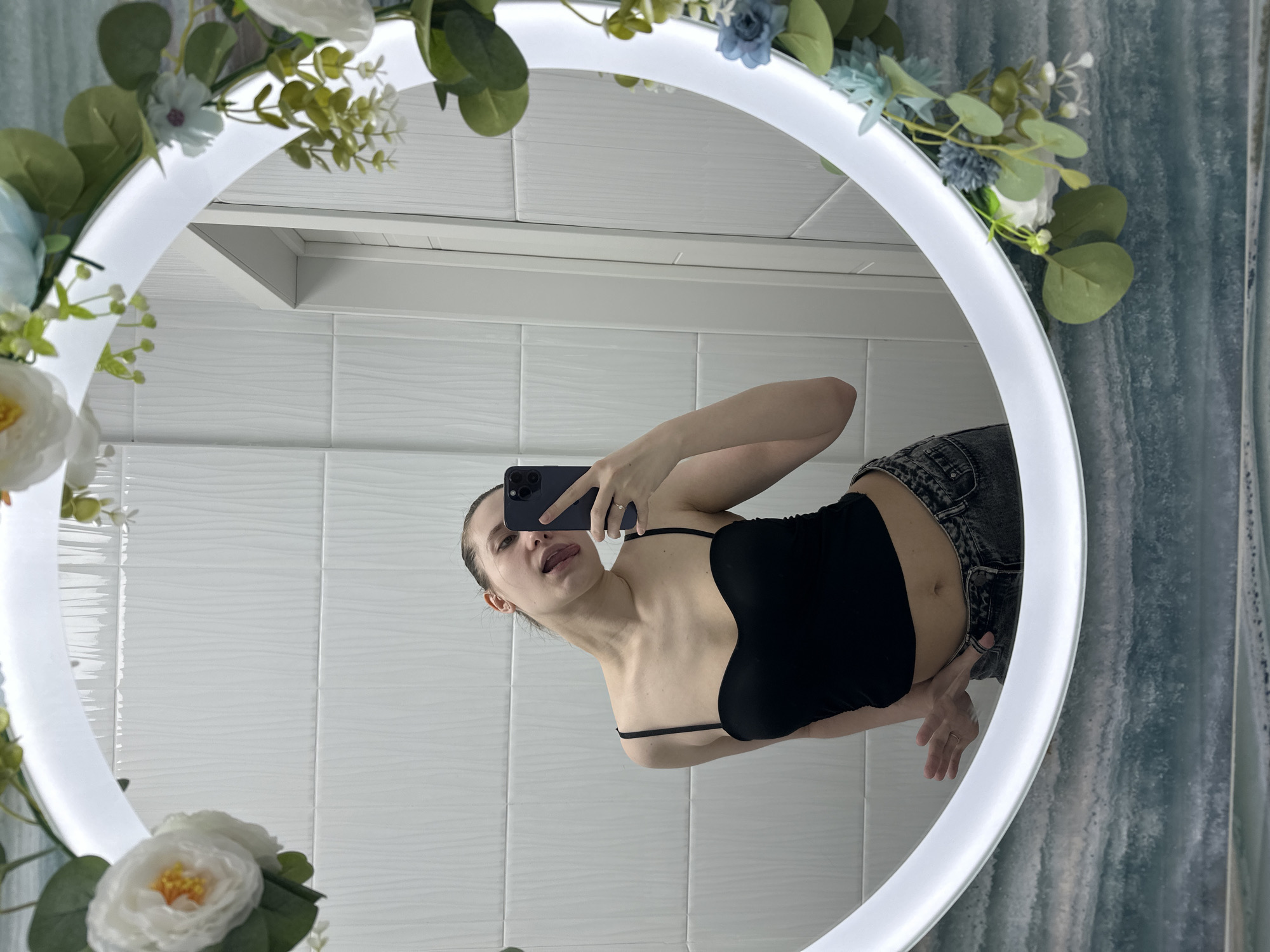 The image displays a mirror selfie taken by a female subject in what appears to be a bathroom or private residence. The subject, a woman with light skin and dark hair pulled back, is visible from the waist up, wearing a black, thin-strapped crop top and dark grey textured jeans. She holds a dark-colored smartphone, consistent with an iPhone, in her left hand, which is used to capture the image. Her head is tilted to the viewer's left, and her tongue is slightly extended. Her right hand rests on her hip. A ring is visible on her left ring finger.
The circular mirror features an illuminated white rim and is decorated with an artificial floral garland comprising white and light blue flowers, along with green foliage. The reflection shows the background behind the subject. The upper wall is covered with white rectangular tiles that have a subtle textured wave pattern. A white door frame is partially visible in the reflection on the right side. Below the mirror, the wall is covered with a blue-green, wavy-patterned tile, resembling a marble or abstract water design.
The location in Moscow, Russia, cannot be determined from the visual information provided in the image.