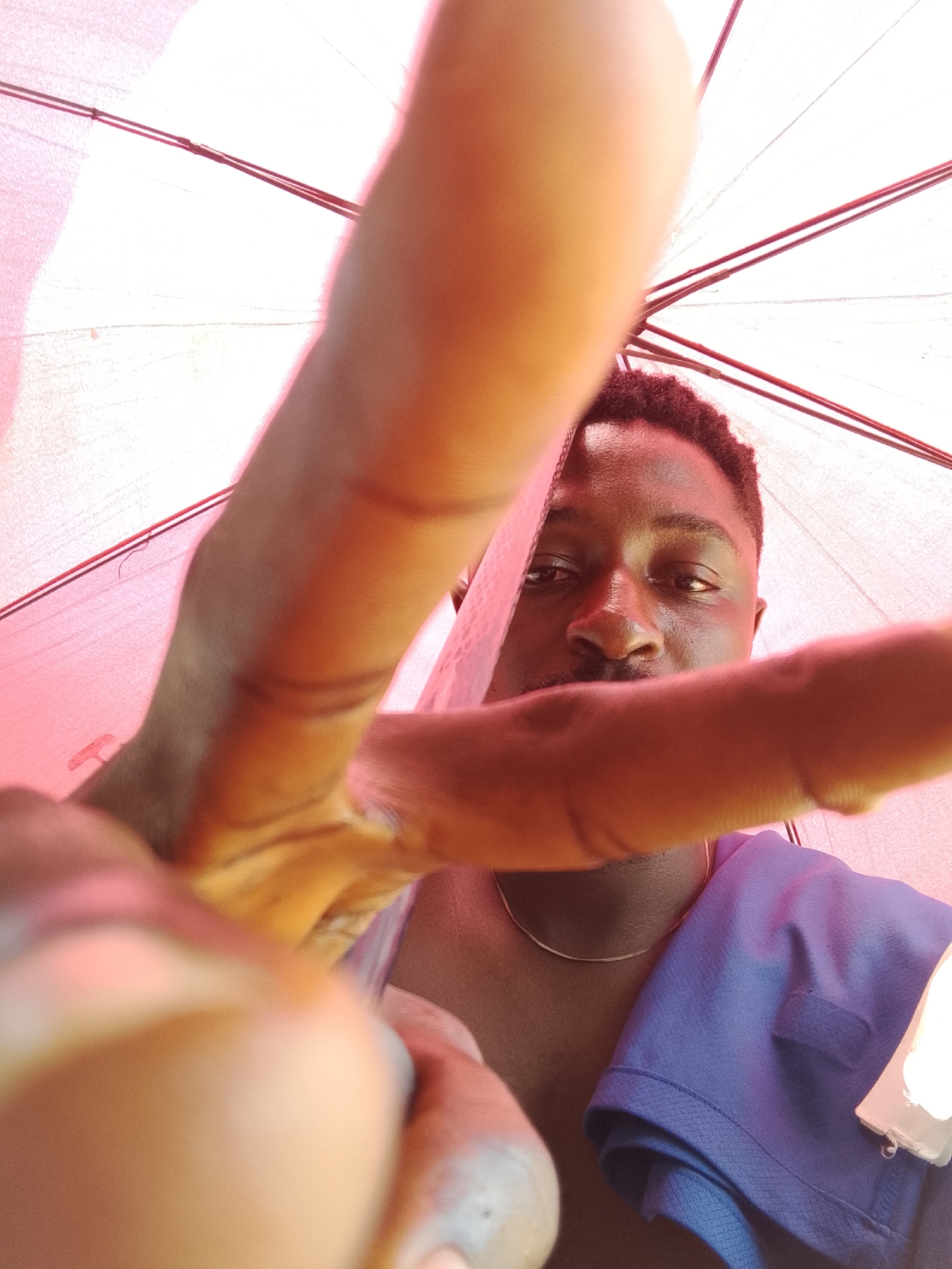 A close-up, low-angle shot features a man with dark skin and short, dark hair, positioned under what appears to be a red and white umbrella. A portion of his face, including his eyes, nose, and upper lip, is visible. He wears a blue shirt and a silver necklace. His fingers are extended towards the camera, obscuring the view. The background is the inside of the umbrella. The location and city are not explicitly deducible from the image.