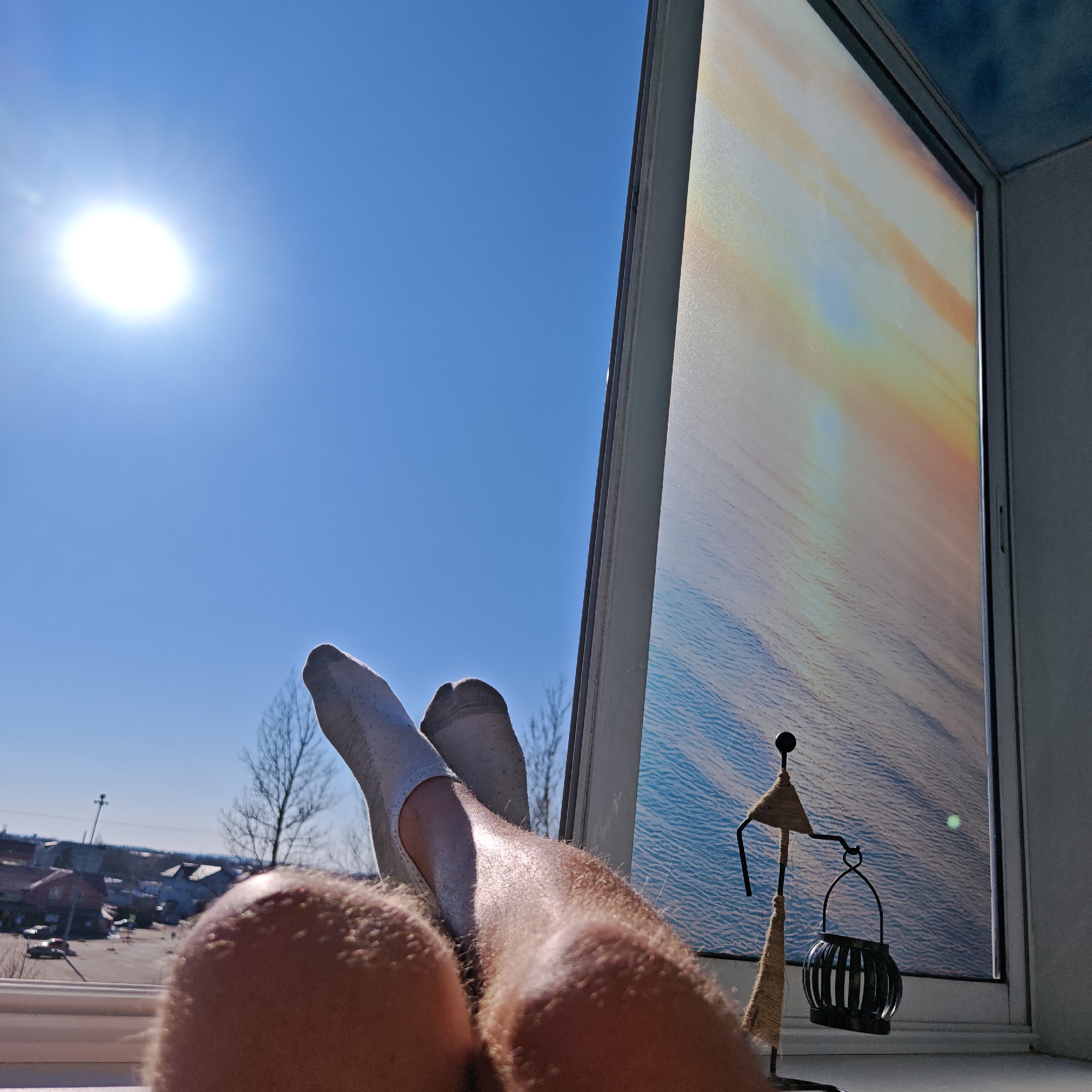 A photograph shows a pair of legs resting on a windowsill inside a building. The feet are covered in low-cut white socks. Outside the window is a clear blue sky with a bright sun, some bare trees, and houses in the distance, with Rostov, Russia buildings being apparent. Inside the window is an ornamental figure of a person holding a lantern-like object. The window has a frosted effect with a water or sky design. The scene is brightly lit with sunlight.