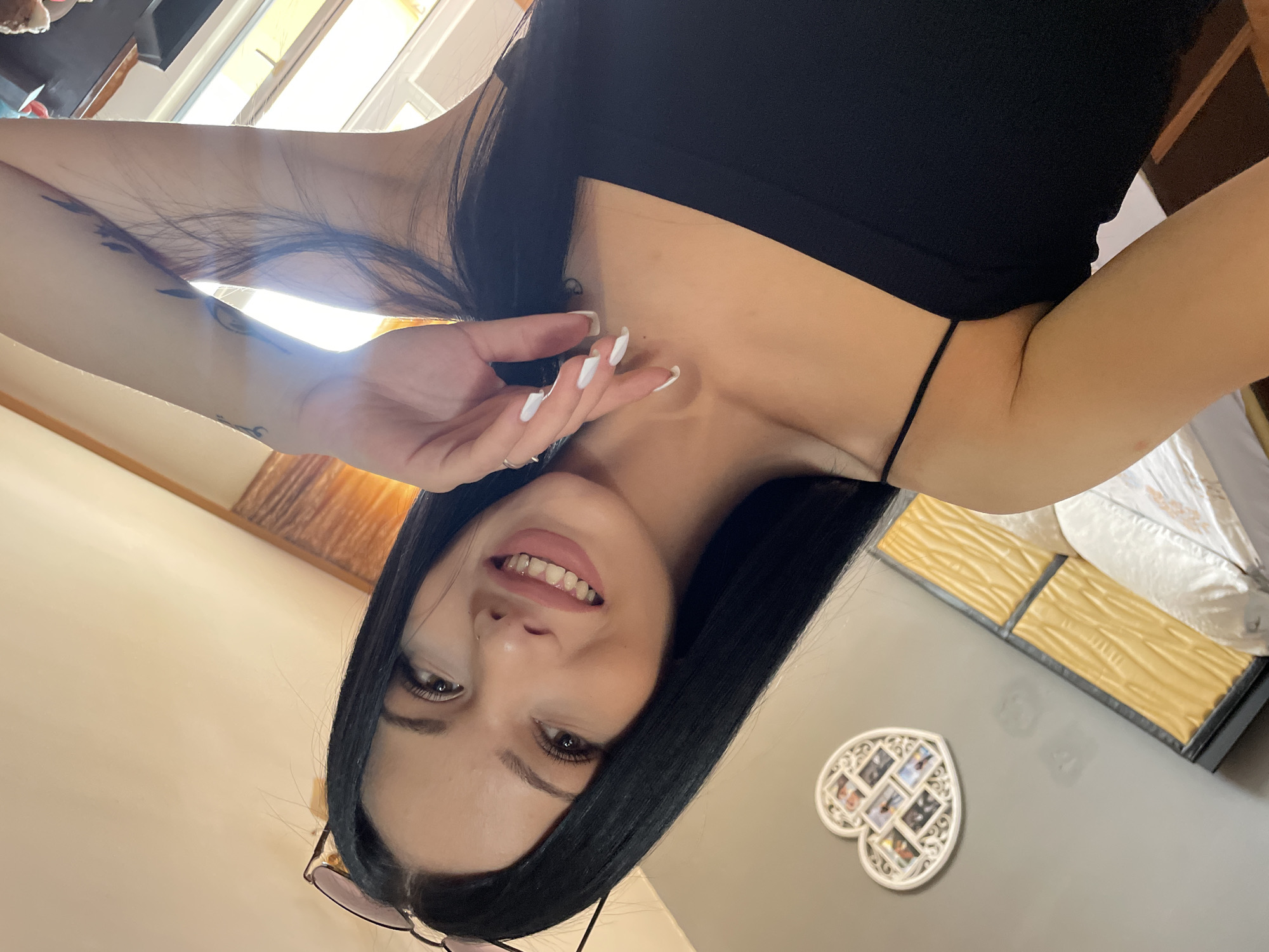 A young woman with long, dark hair and fair skin is taking a selfie in what appears to be a bedroom. She is wearing a black spaghetti strap top and has light pink lipstick on. Her right hand is raised to her face, and her left arm is visible, showing a tattoo on her upper arm. She is smiling, revealing her teeth.
In the background, a grey wall is visible with a heart-shaped photo frame containing several small pictures. Below the frame, a bed with yellow-colored headboard panels and white bedding is partially visible. The room is lit by natural light coming from a window. The overall impression is of a casual, indoor setting.
This image was taken in Stara Zagora, Bulgaria.