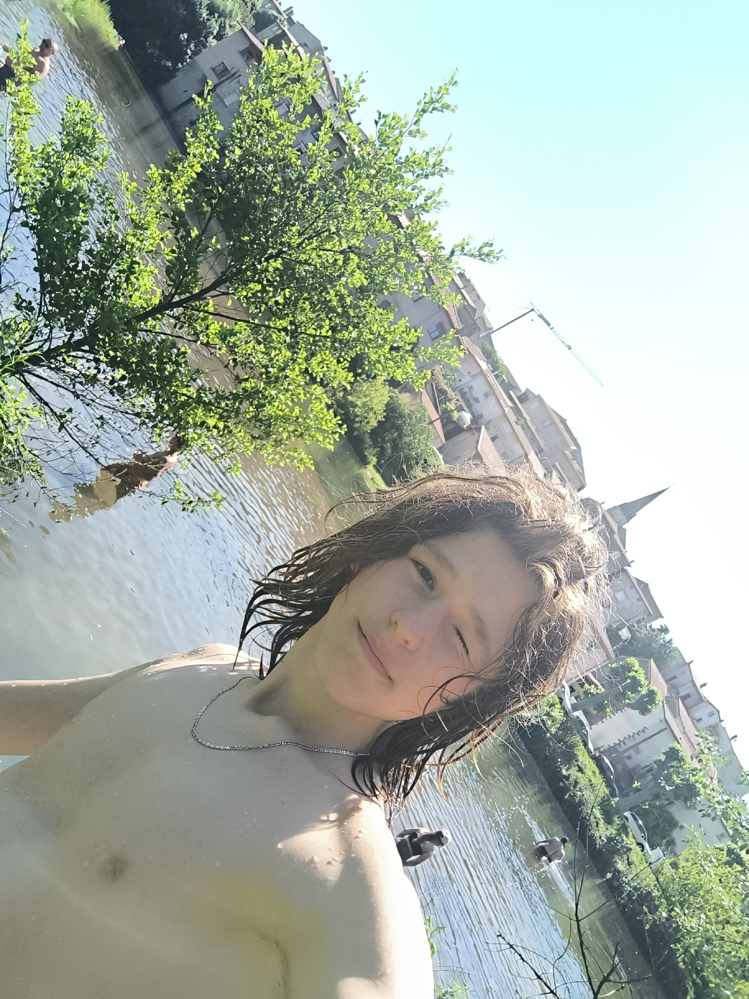 A low-angle, slightly tilted photograph depicts a person in the foreground partially submerged in a body of water, likely a river or large canal. The person is fair-skinned with wet, shoulder-length brown hair, wearing a thin silver necklace, and appears to be either fully nude or wearing minimal clothing. The person is winking and looking directly at the camera.
In the background, the environment includes water, trees, buildings, and other people. The presence of buildings with stone architecture and a church steeple suggests an urban setting. Some people are visible in the water behind the subject.
The location is Chézenas, France, based on identifiable architectural elements and the landscape.
