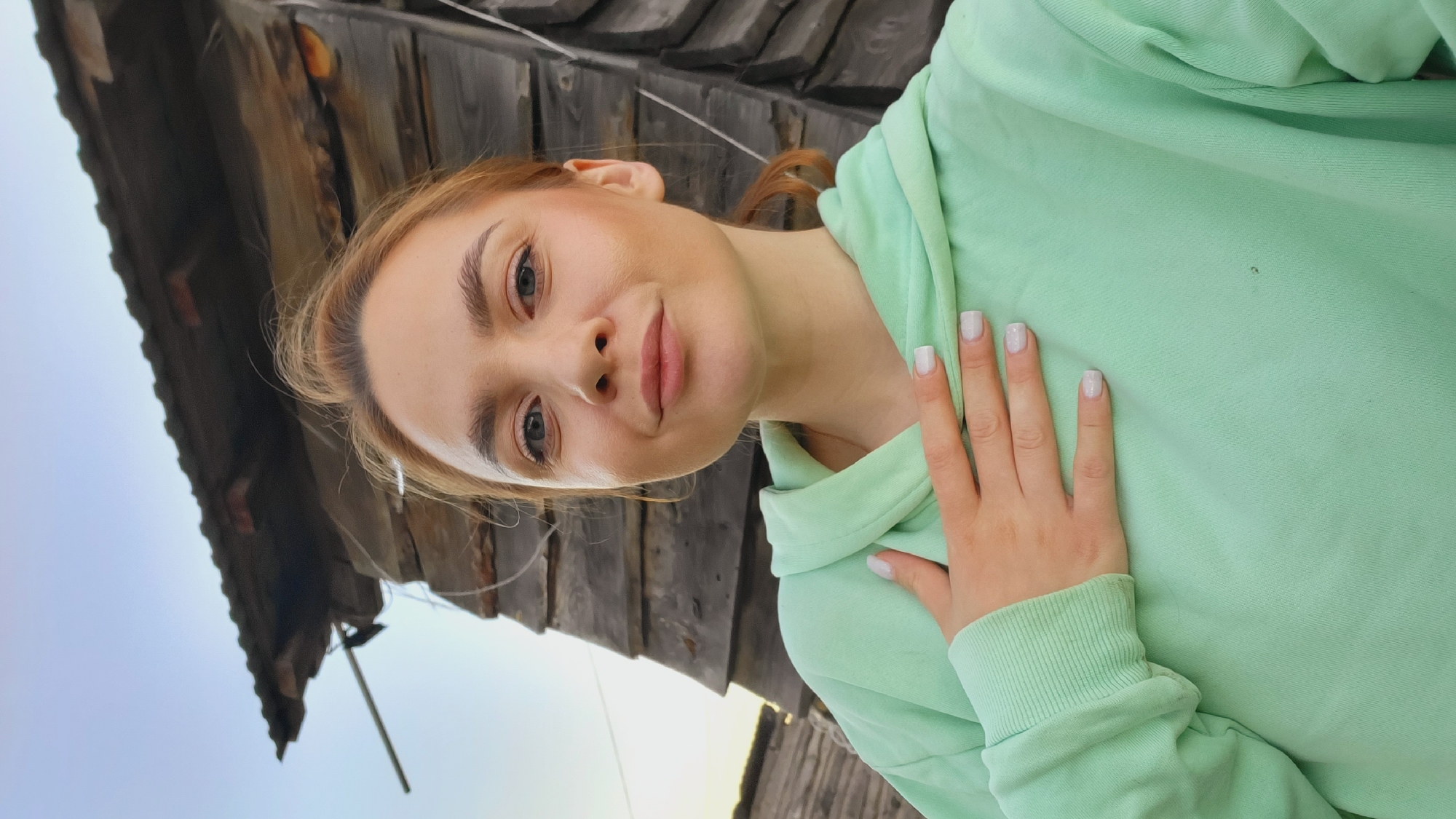A selfie of a young fair-skinned woman with reddish-blonde hair wearing a mint green hooded sweatshirt. Her right hand is placed on her chest. The background appears to be an old, wooden structure with a grey sky visible behind it. The location is Зипуново, Russia.