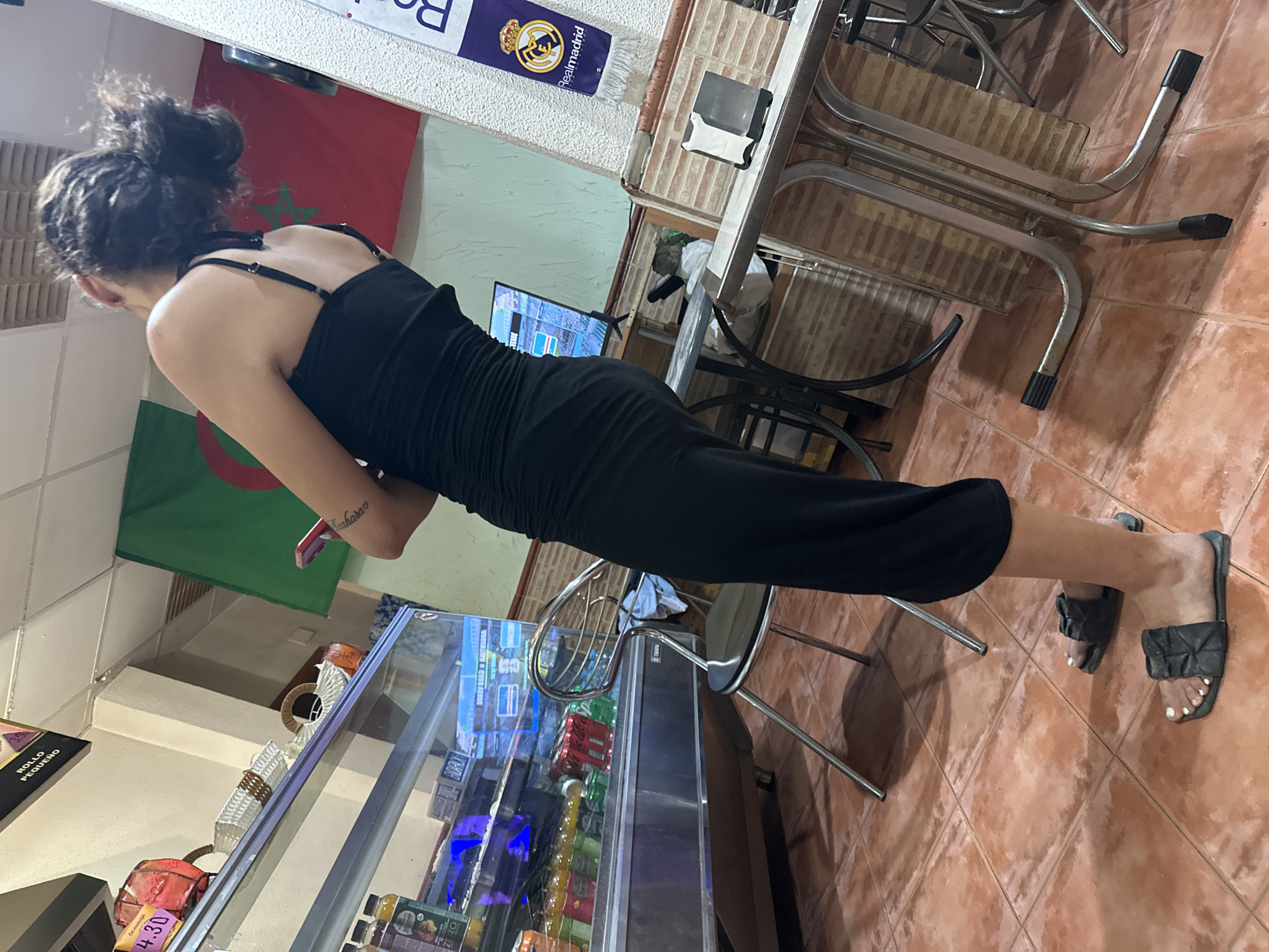In an interior setting that appears to be a casual dining establishment or a cafe in Alicante, Spain, a woman with dark hair pulled up in a bun stands facing away from the camera. She is wearing a ruched black dress and black slide sandals. Her left arm is bent, and she is holding a red smartphone. In the background, a refrigerated display case contains beverages. Two flags are visible on the wall: one is green with a red star, and the other is red with a white emblem. A Real Madrid banner is also displayed. The flooring is tiled in a terra cotta color. The lighting suggests an indoor commercial space.