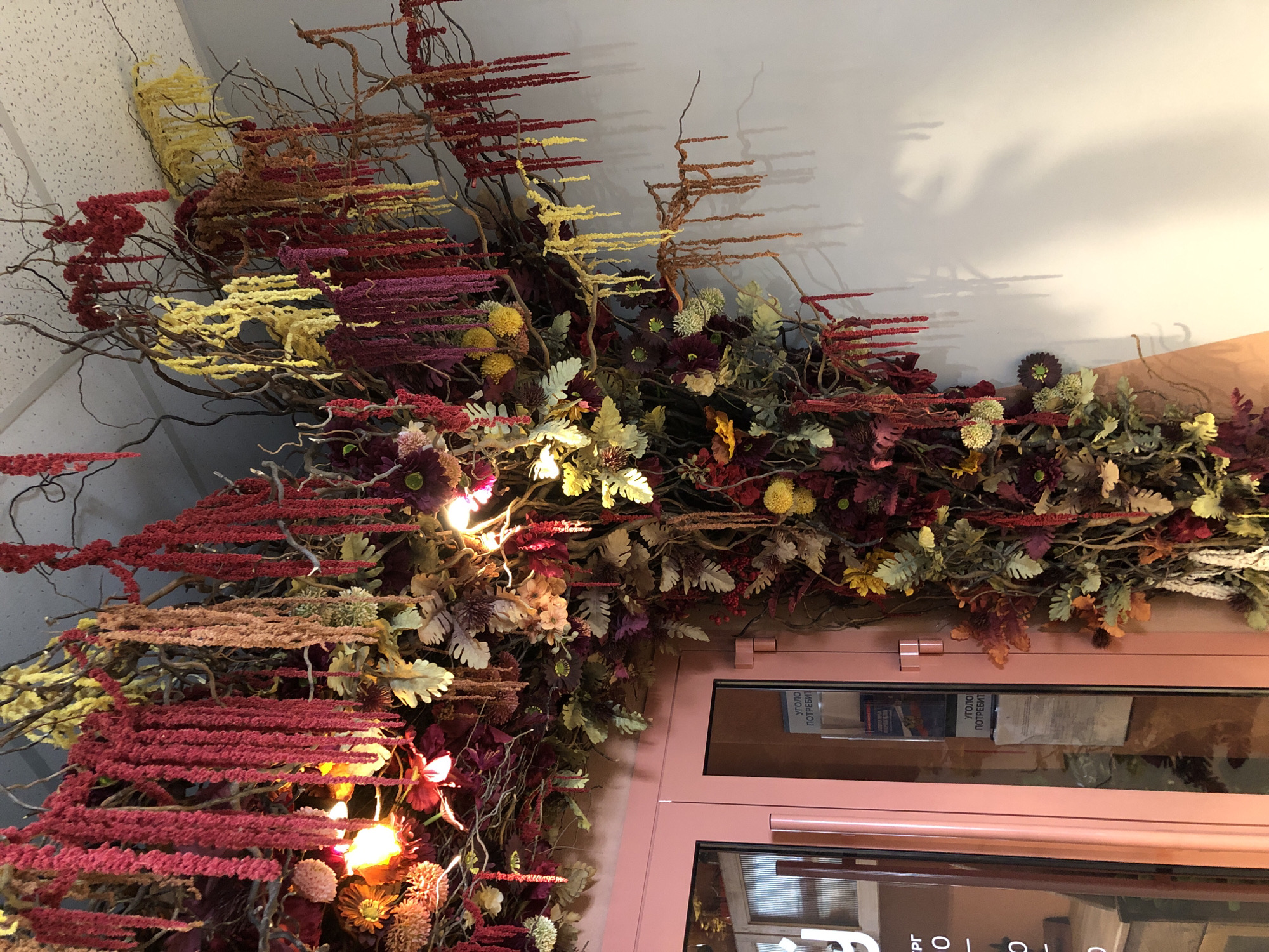 An indoor scene within a commercial establishment in Chaikovsky, Russia, features an elaborate decorative installation of dried or artificial flora. The arrangement occupies the upper left quadrant of the image, extending across the ceiling and down the right wall. It is composed of numerous bare tree branches intertwined with dense clusters of red, yellow, and purple cascading floral elements, along with various types of foliage and rounded blooms. Several integrated warm-toned lights illuminate portions of the display. To the left, a set of double-leaf doors with pastel pink frames and glass panels are visible, one featuring a white sign with Cyrillic text, including "УГОЛОК ПОТРЕБИТЕЛЯ" (Consumer Corner). The ceiling is a white acoustic tile grid. The adjacent wall to the right is plain white, exhibiting distinct shadows cast by the floral arrangement. No individuals are present.