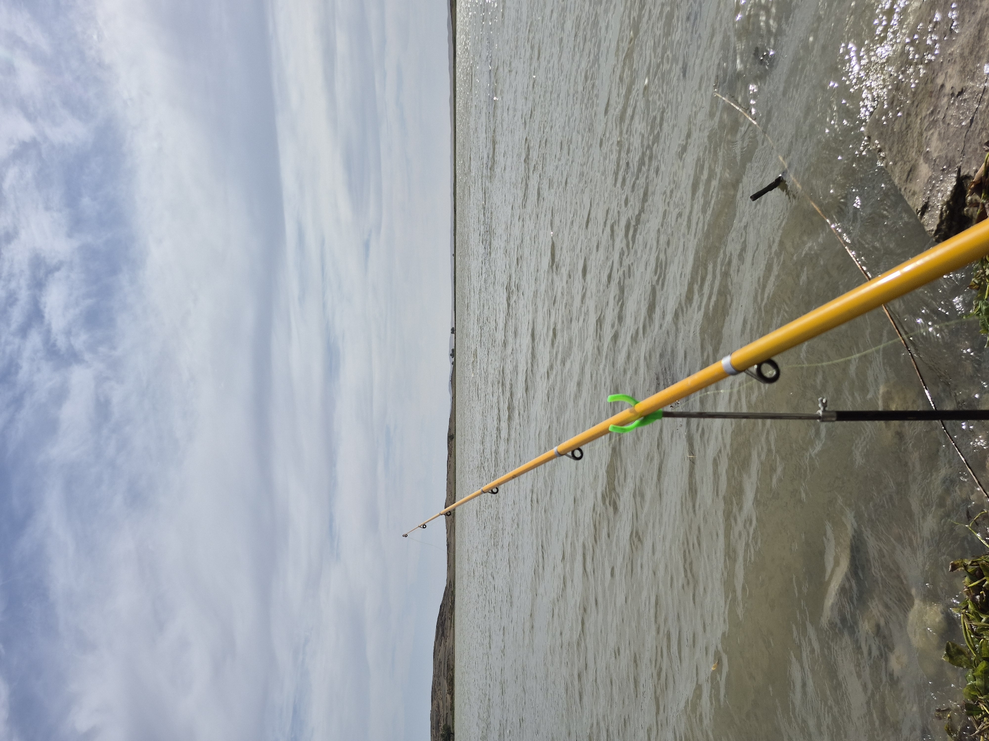 The image shows a yellow fishing rod resting on a rod holder near the edge of a body of water. The water is calm with small ripples, and the bottom is visible in the shallow areas. The shore is rocky with some green vegetation. In the background, there is a low, hilly landscape under a cloudy sky. The location is identified as Көктіңкөлі, Kazakhstan. There are no people or animals visible in the image.