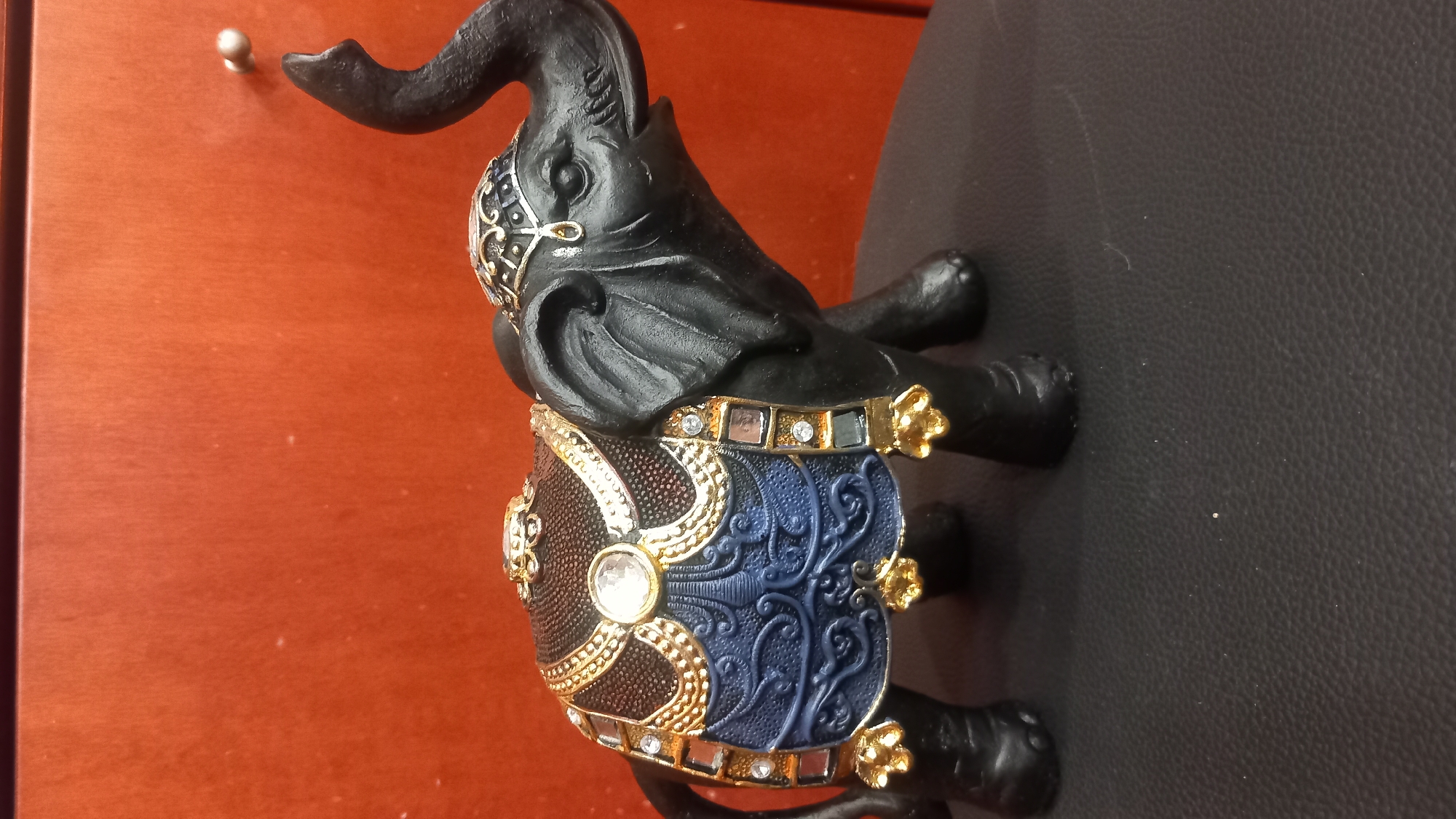 A black elephant figurine is displayed on a dark surface, likely a piece of furniture. The figurine is adorned with a richly decorated saddle featuring blue and gold accents, intricate patterns, and sparkling embellishments that resemble gems and metallic threads. The elephant's trunk is raised in the air, and its head is turned to the right. The background is a warm, reddish-orange surface with a subtle wood grain texture. There are no people or animals present in the image, and no specific landmarks or environmental features indicate a location within the city of Kos, Greece.