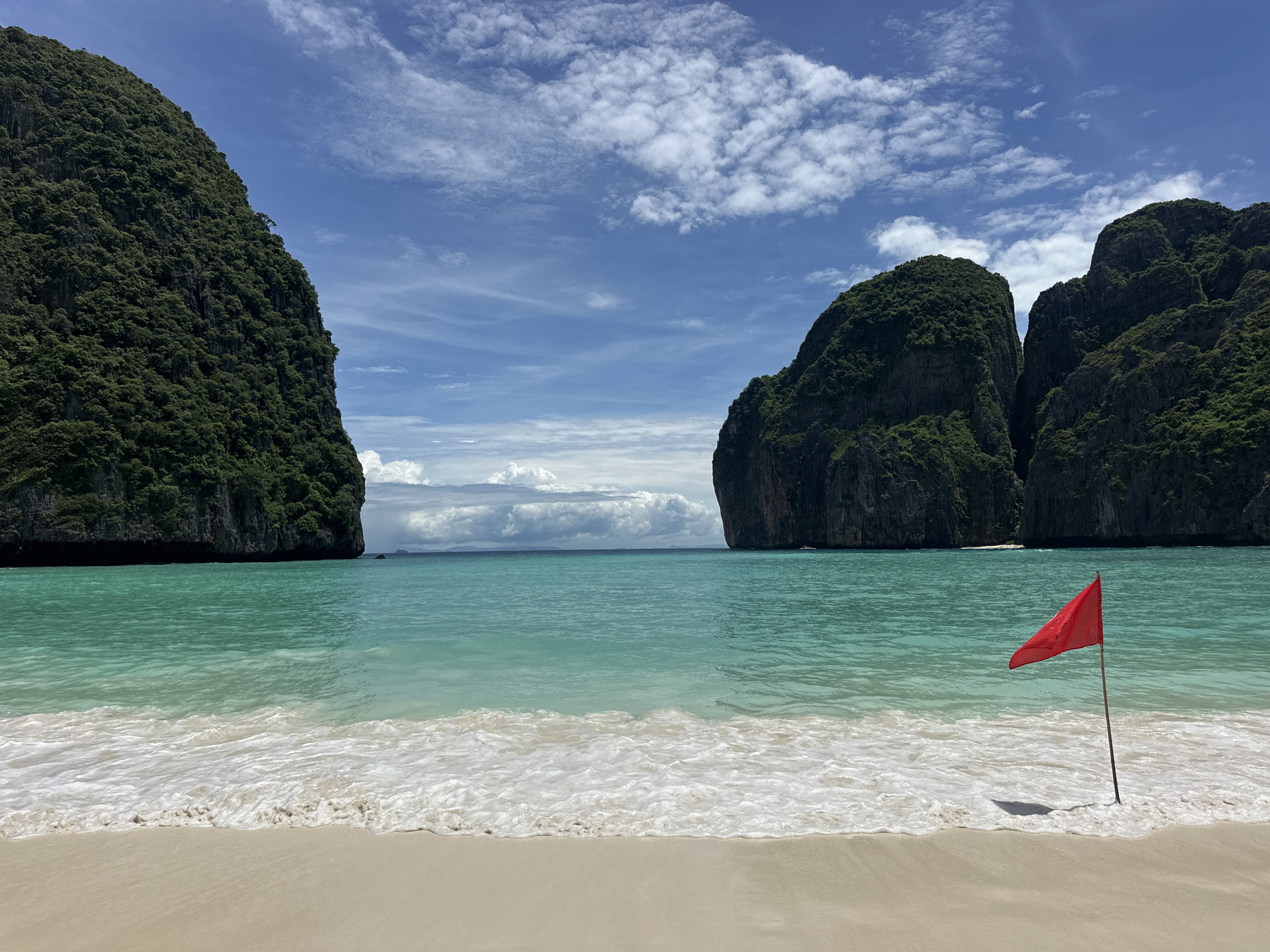 The image depicts a serene tropical beach scene with turquoise waters lapping onto a sandy shore. Towering, verdant limestone cliffs flank the bay, creating a dramatic backdrop against a bright blue sky dotted with fluffy white clouds. A lone red flag stands planted in the sand near the water's edge, its fabric gently fluttering in the breeze. The scene evokes a sense of tranquility and natural beauty. This location is Maya Bay, on Koh Phi Phi Leh island, which is a popular tourist destination accessible from Ao Nang, Thailand.