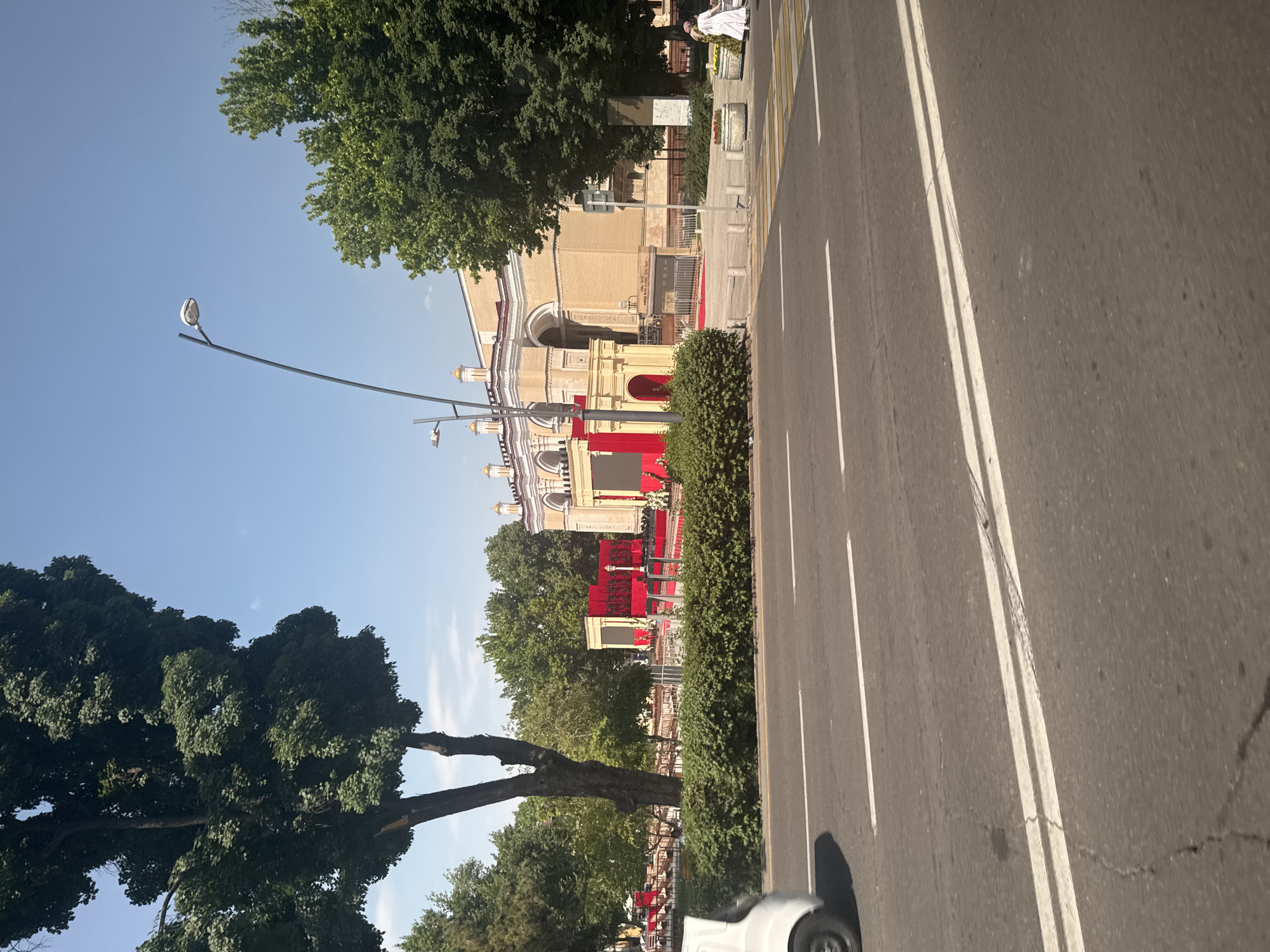 The image depicts a street scene in Tashkent, Uzbekistan, oriented sideways. A multi-lane road with white lines runs from the bottom right to the top right. A building, seemingly a government or cultural institution, is visible on the left side of the road. The building is decorated with red fabric or flags, suggesting an event or ceremony. Trees line the road, and the sky is blue. A lamppost stands near the building. A white car is partially visible in the bottom right corner of the image.
