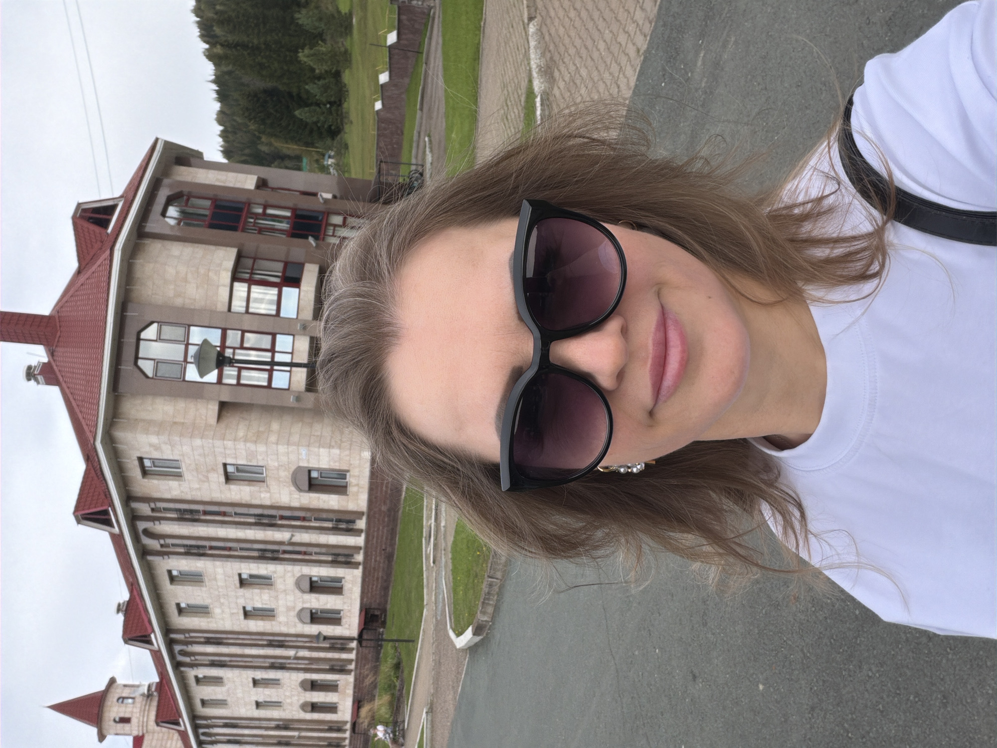 A woman with shoulder-length brown hair and sunglasses stands in front of a light-colored, multi-story building with a red roof, possibly a hotel or administrative building. The building has arched windows and decorative vertical elements. Behind the building, a dense forest of green trees covers a sloping hillside. The woman is wearing a white t-shirt and a black bag strap is visible over her right shoulder. The scene appears to be outdoors on a paved area, possibly a road or walkway, with some grass and a low wall visible in the background. The sky is overcast. This location is identified as being in the city of Assy, Russia.