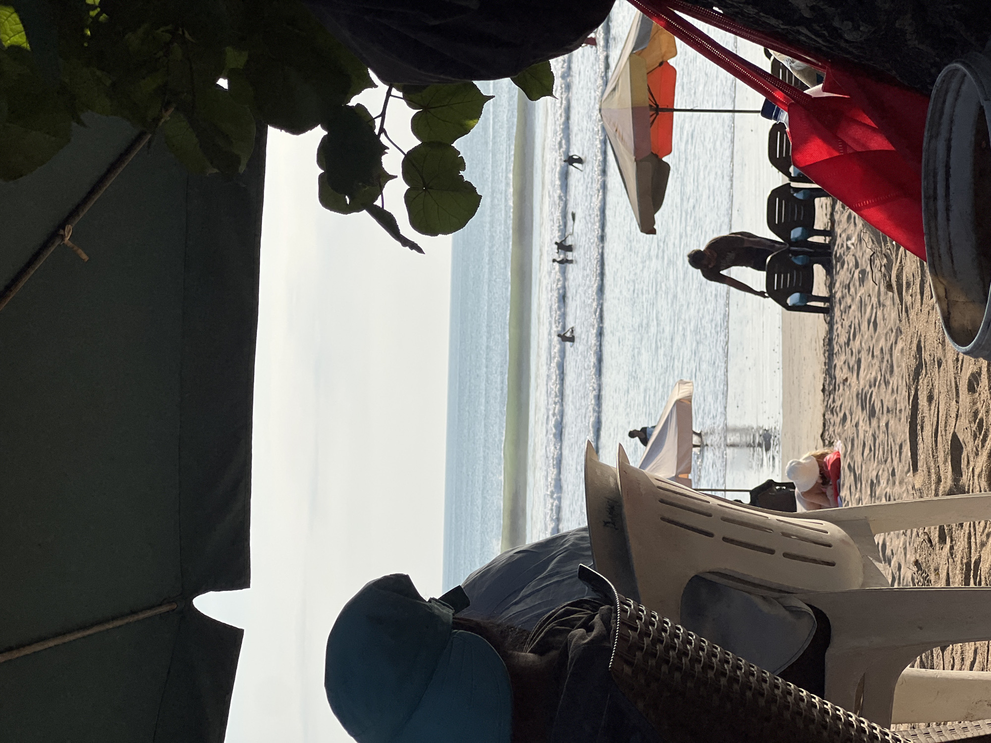 This is a photograph of Kuta Beach in Bali, Indonesia. The image is taken from a shaded area, likely under an umbrella or canopy, looking out towards the ocean. In the foreground, a person wearing a light blue hat is seated, facing away from the camera, their back to the viewer. Next to them, a woven chair is visible. Further into the scene, the sandy beach is dotted with several beach chairs and umbrellas, some occupied by people. The ocean is calm with gentle waves, and several individuals are visible in the water, some appearing to be surfing. The overall impression is a relaxed beach scene on a sunny day.