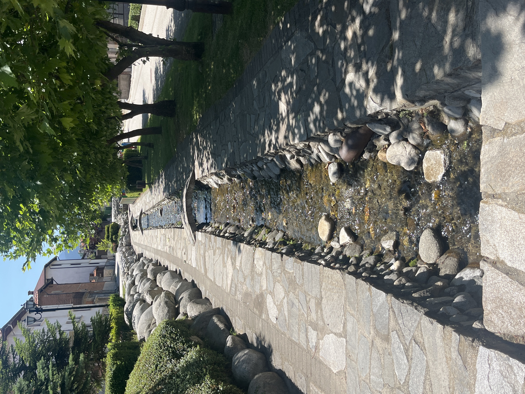 The image depicts a charming, tree-lined street in Bansko, Bulgaria, featuring a shallow, clear stream running through the center. The stream is bordered by stone pathways and large, rounded rocks. Small, arched bridges cross the stream at intervals. On the left side of the stream, there are manicured bushes and a stone wall made of large, smooth boulders. Buildings with white and brick facades are visible in the background, suggesting a residential or commercial area. On the right side, a row of mature trees with lush green foliage casts dappled shadows on the pathway. The scene is bathed in bright sunlight, indicating a clear, pleasant day. There are no people or animals visible in the frame.