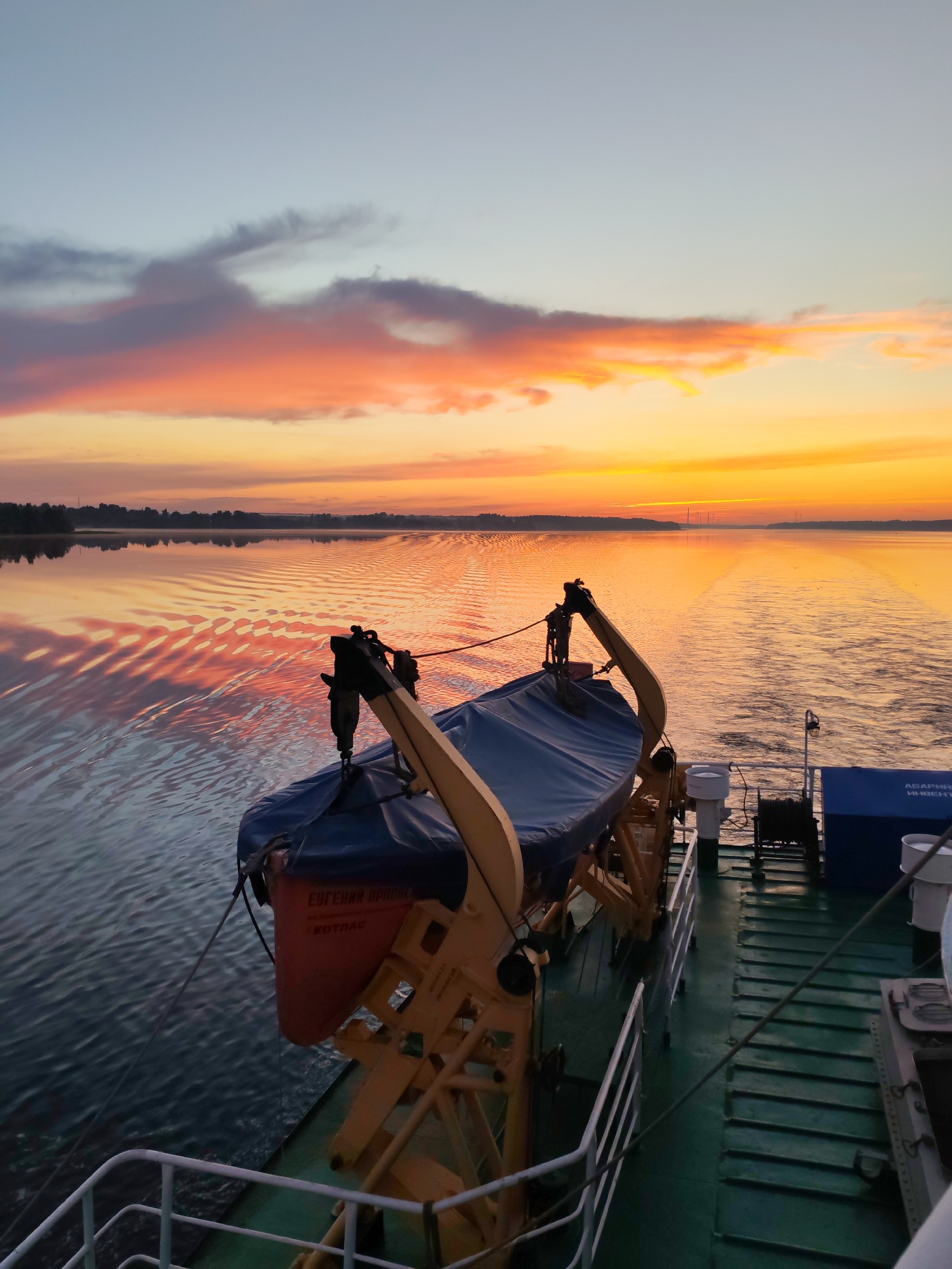 The image captures a sunrise over a body of water, as seen from the deck of a ship. The sky is painted with vibrant hues of orange, pink, and yellow, with some wispy clouds. The water reflects the colors of the sky, creating a shimmering effect. In the foreground, a lifeboat covered with a blue tarp is secured to the deck by a davit. The deck itself is green, with metal railings and various pieces of equipment, including a winch and a blue storage container. The overall scene conveys a sense of calm and the beginning of a new day.