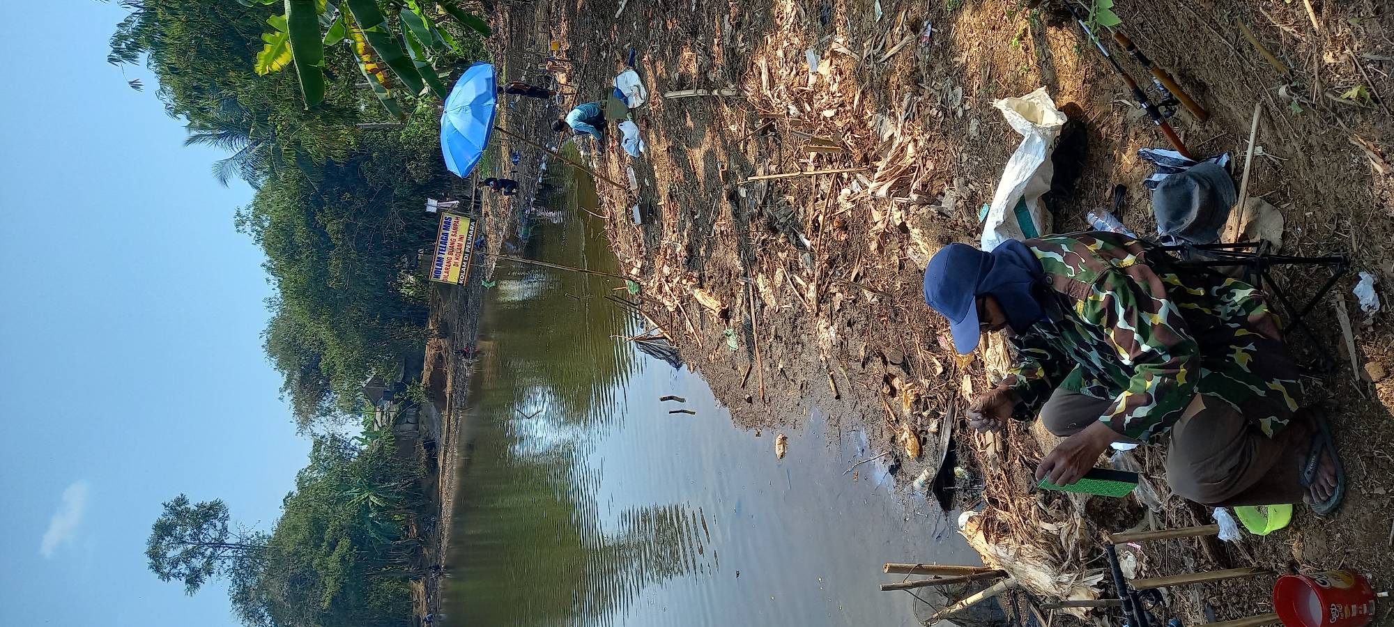 In Sidamulya, Indonesia, a man in camouflage clothing and a blue cap is sitting on a folding chair by a body of water. He is facing the water and appears to be preparing fishing gear. There are fishing rods set up in the ground around him, as well as a small yellow bucket and a larger red bucket. The ground is covered in dirt, dry leaves, and some scattered trash. In the background, other people can be seen fishing by the water. A bright blue umbrella is set up to provide shade. Lush green trees line the opposite bank of the water, and a sign in Indonesian is visible in the distance. The sky is clear and blue.