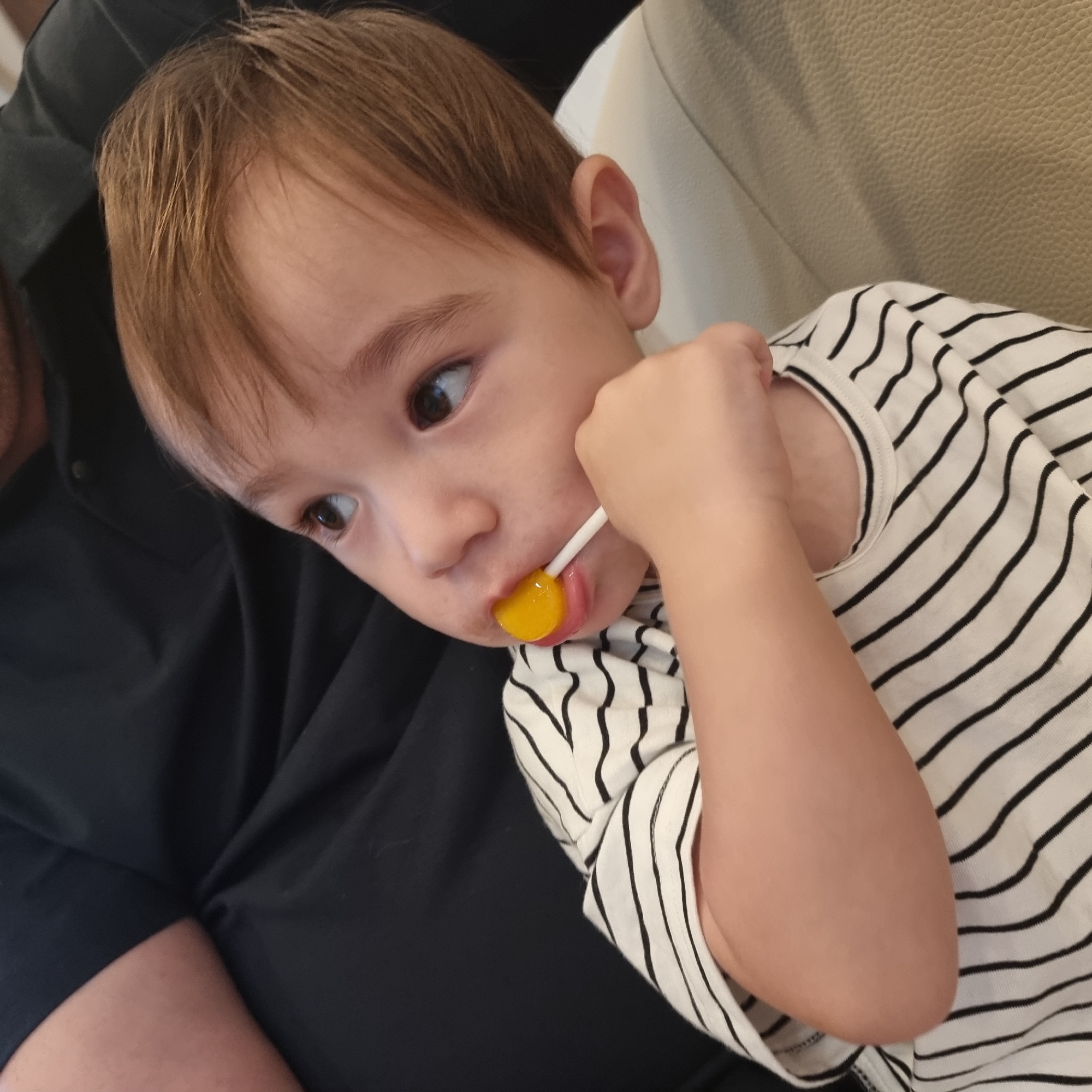 In an indoor setting, a young child with light brown hair is pictured, wearing a black and white striped t-shirt. The child is looking upwards and to the right, with a yellow lollipop in their mouth. Their right hand is near their mouth, holding the lollipop stick. The child appears to be sitting on someone's lap, indicated by the presence of a dark fabric, possibly clothing, behind and to the left of the child, and a lighter colored textured surface, likely furniture, to the right. The lighting suggests an ambient indoor environment. The image does not provide any specific landmarks or features to determine its location within Wonju-si, South Korea.