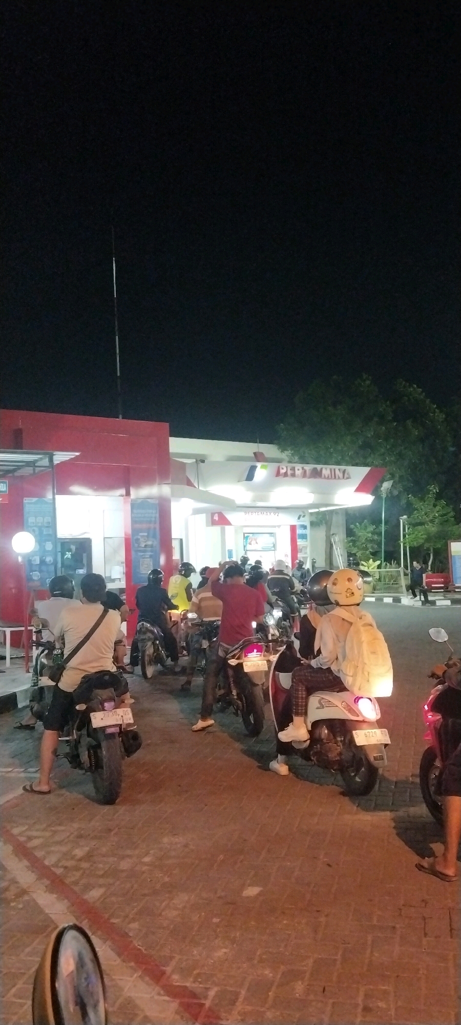 The image depicts a nighttime scene at a Pertamina gas station in Karangturi, Indonesia. A queue of motorcyclists is waiting for fuel. In the foreground, a person on a scooter wearing a light-colored hoodie and plaid pants is visible, with a white backpack on the rear seat. Other motorcyclists in the queue are wearing helmets and various casual clothing. The gas station itself is illuminated, with the "PERTAMINA" logo clearly visible on the canopy. Trees are present in the background, silhouetted against the dark night sky. The ground is paved with interlocking bricks. The overall impression is one of routine activity at a public service station during the evening hours.
