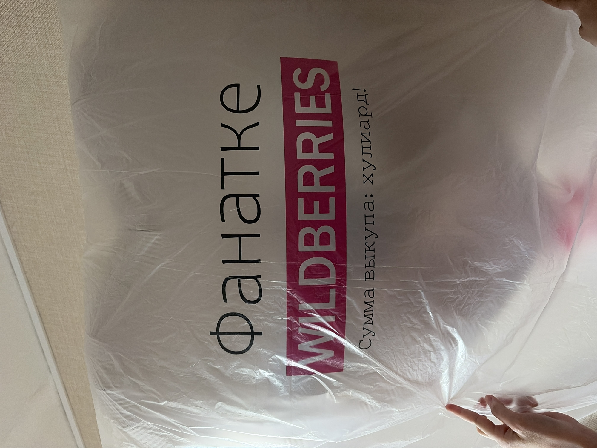 A partially obscured photograph depicts a product package on a textured surface, possibly a bed or sofa. The package, made of clear plastic, features the "Wildberries" logo prominently displayed. The text "фанатке" is printed to the left of "Wildberries," and the phrase "Сумма Выкупа: хулиард!" is visible to the right. A person's hand is visible at the bottom of the frame, seemingly manipulating the plastic packaging. The scene is indoors, and the photograph's composition suggests a close-up view. The precise location within the city of Chaikovsky, Russia, cannot be determined from the visual information provided.