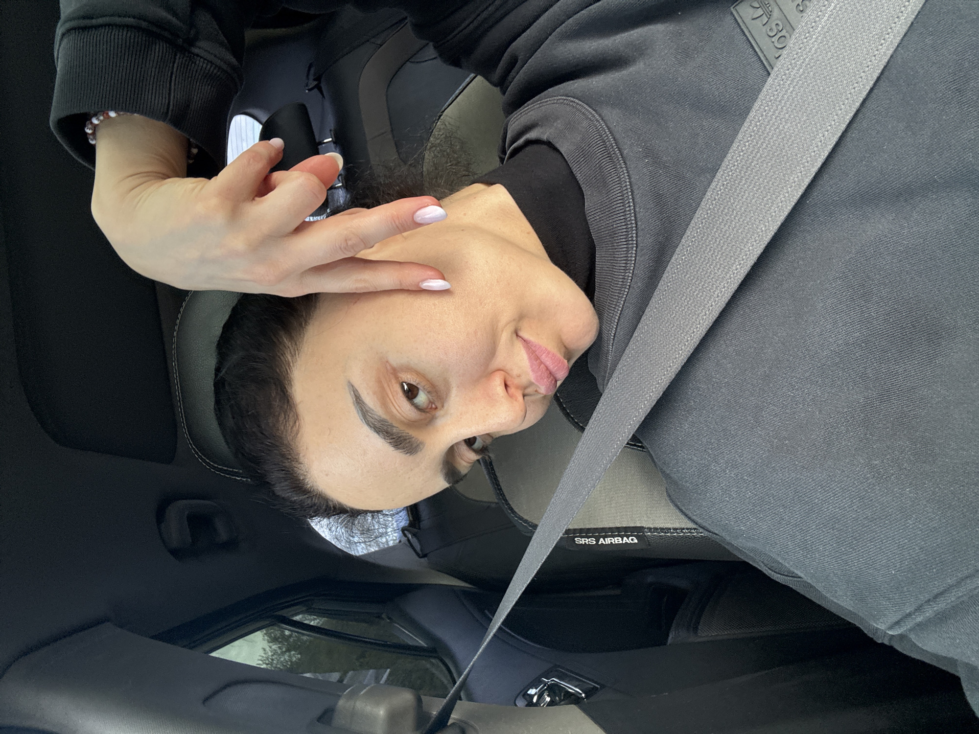 A woman is in the driver's seat of a vehicle, wearing a gray sweatshirt and a seatbelt. She is making a hand gesture towards her face. The interior of the car is black and gray. The scene is likely located inside a vehicle, but the city of Tbilisi, Georgia, is not discernible from the image.