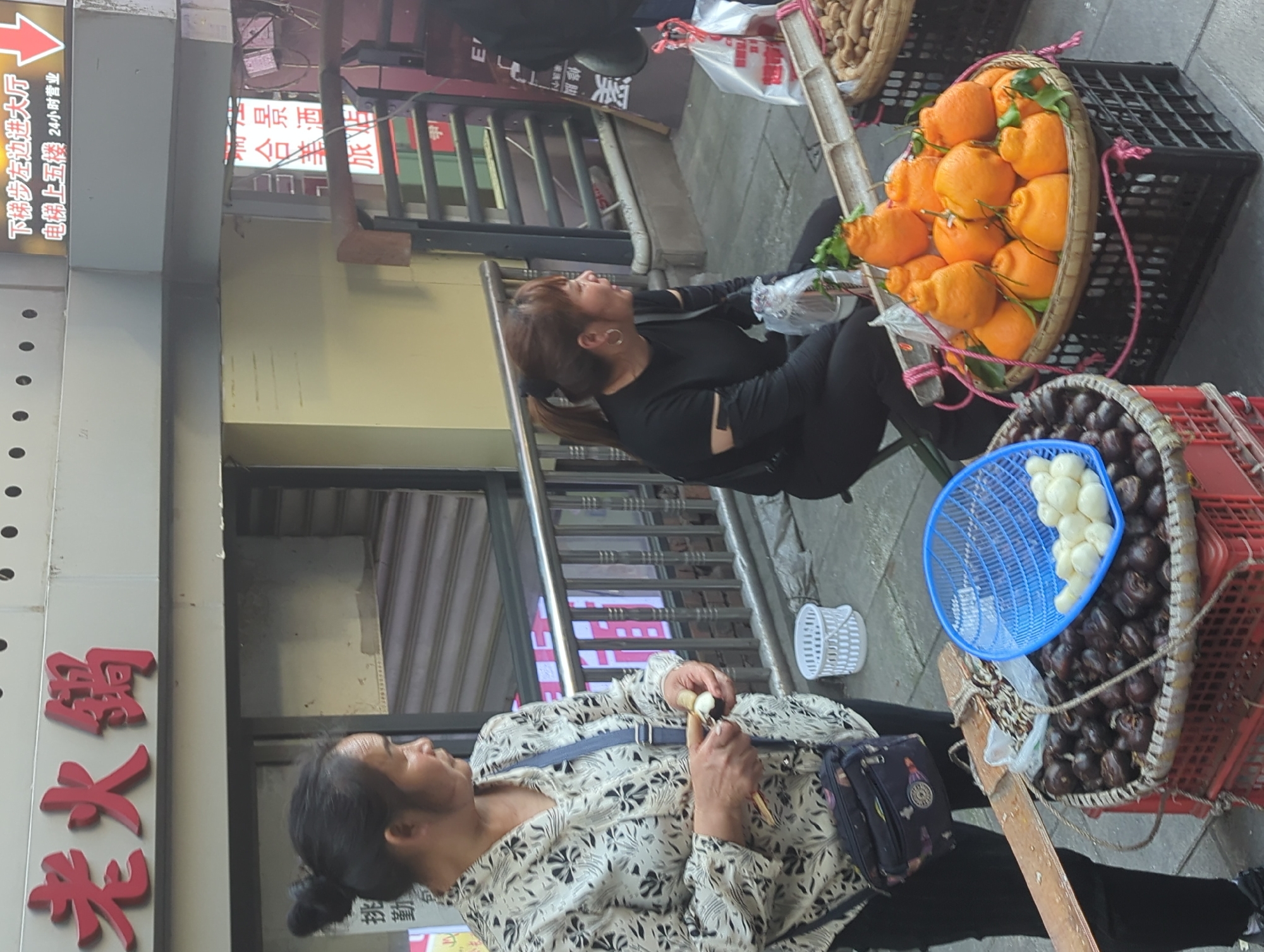 This image depicts a bustling street scene, likely a market, in an urban area during daytime. The perspective is tilted, showing the ground and various stands alongside building facades.
Two women are visible. In the foreground, an older woman with her hair tied back wears a light-colored, patterned long-sleeved shirt and black pants, with a black shoulder bag. She is looking to her left and appears to be in the process of peeling a small, white, ovoid item, possibly a water chestnut, using a small tool. Behind her, a younger woman with reddish-brown hair tied in a ponytail is seated. She wears a black long-sleeved top and black pants, looking upwards and to her right, seemingly resting or observing.
Next to the younger woman, there are several street vendor stands. One prominent display features a large basket filled with numerous bright orange, bumpy-skinned citrus fruits, resembling 'ugly fruit' or Dekopon, arranged in a large, flat wicker basket. Below this, another basket contains a large quantity of dark, round items, most likely water chestnuts, and a blue plastic colander holding a pile of the same items, peeled and white.
The background shows building exteriors with windows and metal railings, suggesting multi-story structures. Various signs with Chinese characters are mounted on the buildings. Visible text includes "老火锅" (Old Hotpot), "下梯步左边进大厅 电梯上五楼 24小时营业" (Go down the steps on the left to enter the hall, elevator to the fifth floor, open 24 hours), "三合景" (likely part of a business name), and "修脚" (pedicure). The scene captures a moment of daily life and commerce on a street in Yuzhong District, China.