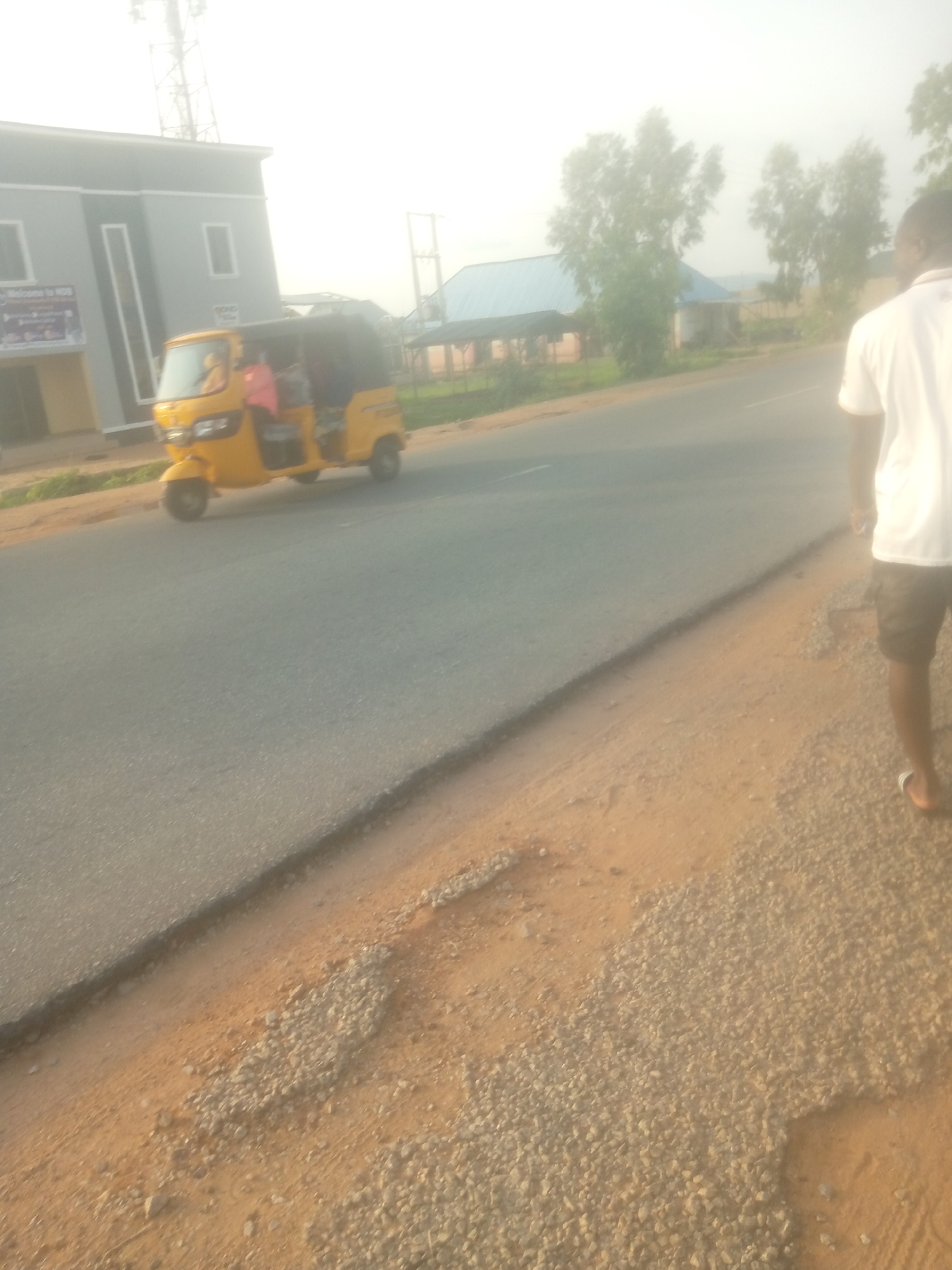 A yellow auto-rickshaw, carrying several passengers, travels along a paved road in Jalingo, Nigeria. The auto-rickshaw is positioned to the left of the frame, with its front facing slightly towards the viewer. The passengers appear to be seated inside the enclosed cabin of the vehicle. To the right of the auto-rickshaw, a person is walking away from the camera, also on the paved road. This individual is wearing a white t-shirt and dark shorts. The environment is characterized by a road with a gravel shoulder. In the background, there are buildings, trees, and a cell tower, indicating an urban or semi-urban setting. The sky is overcast, suggesting a hazy or misty atmospheric condition. The scene appears to be captured during daylight.
