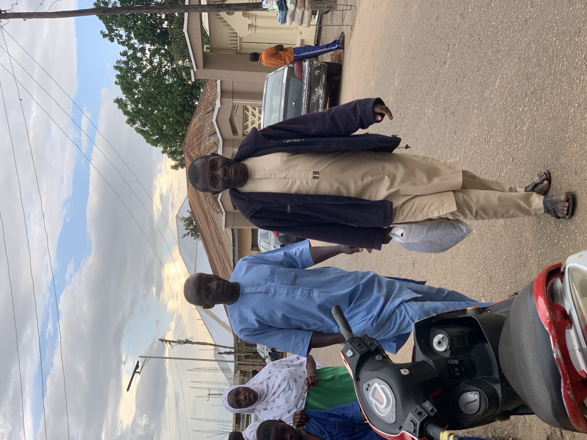 In Jos, Nigeria, two men walk down a paved street, with one man on the right smiling at the camera and wearing a beige tunic, pants, and a dark blue jacket. The other man, on the left, is wearing a light blue tunic and pants. Behind them, a woman in a white headscarf and green skirt walks with a child. A red and black scooter is parked on the left side of the street. In the background, several buildings with corrugated metal roofs are visible, along with trees and utility poles. The sky is partly cloudy.