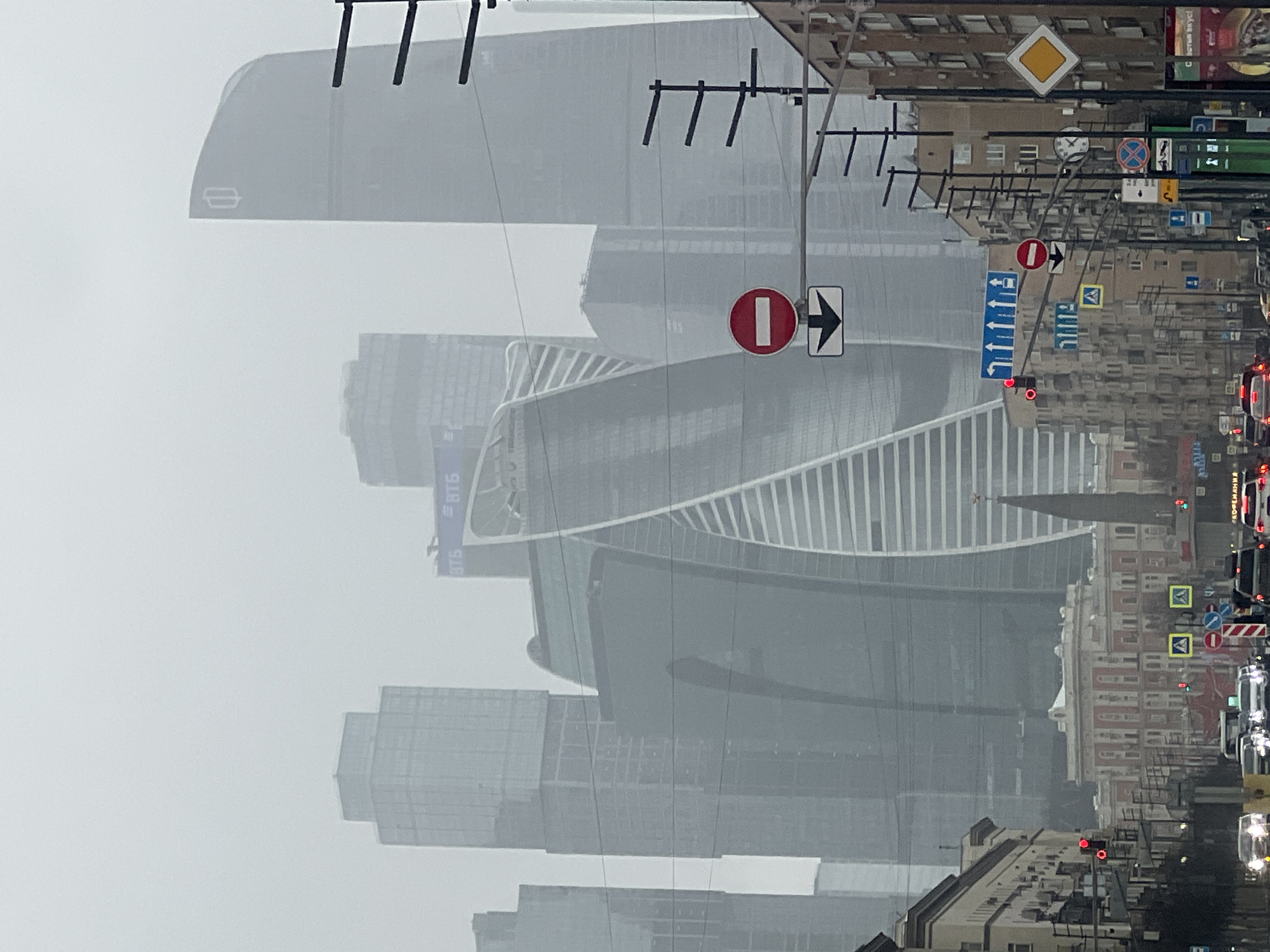 The image depicts a street scene in Moscow, Russia, characterized by heavy fog. The foreground features a series of traffic signs, including "No Entry" and directional arrows. Several high-rise buildings are visible in the background, obscured by the fog. These skyscrapers are part of the Moscow International Business Center, also known as Moscow City. The architecture is modern, with many buildings featuring glass facades. Along the street are traditional building with windows. The sky is overcast, contributing to the overall foggy atmosphere. Cars are driving on the street.